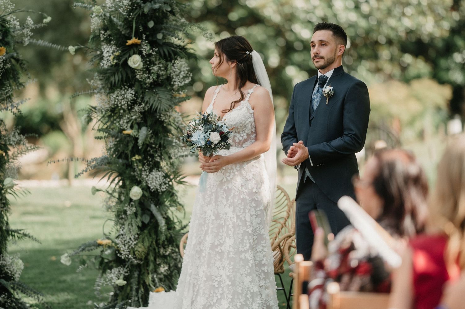 Outdoor wedding ceremony with floral arch and guests seated on wooden chairs.