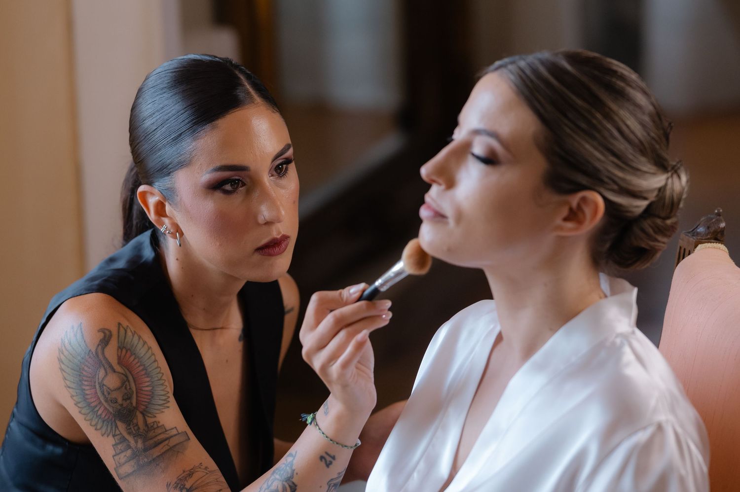 A makeup artist with tattoos applies lipstick to a client wearing a white robe during a beauty session.