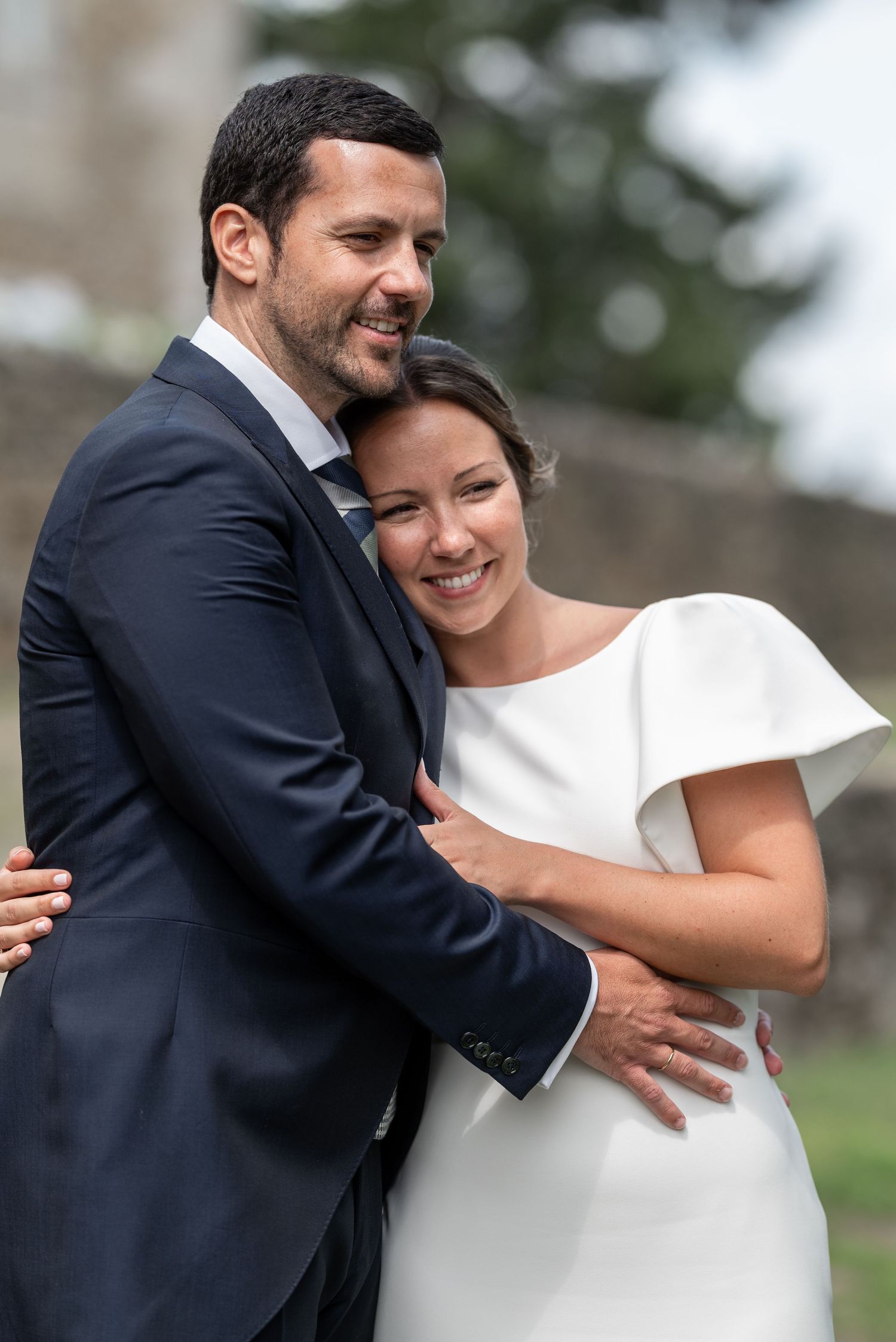 A joyful wedding couple embraces outdoors, with the bride wearing a modern white dress and groom in navy suit.