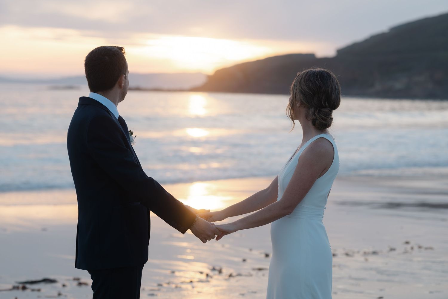A couple in wedding attire holds hands while walking along a beach at sunset with mountains in the background.