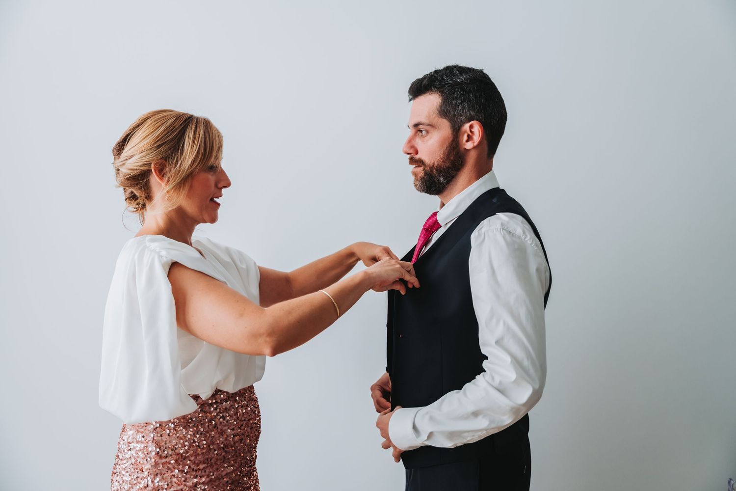 A couple in formal attire share an intimate moment while adjusting their wedding day attire.