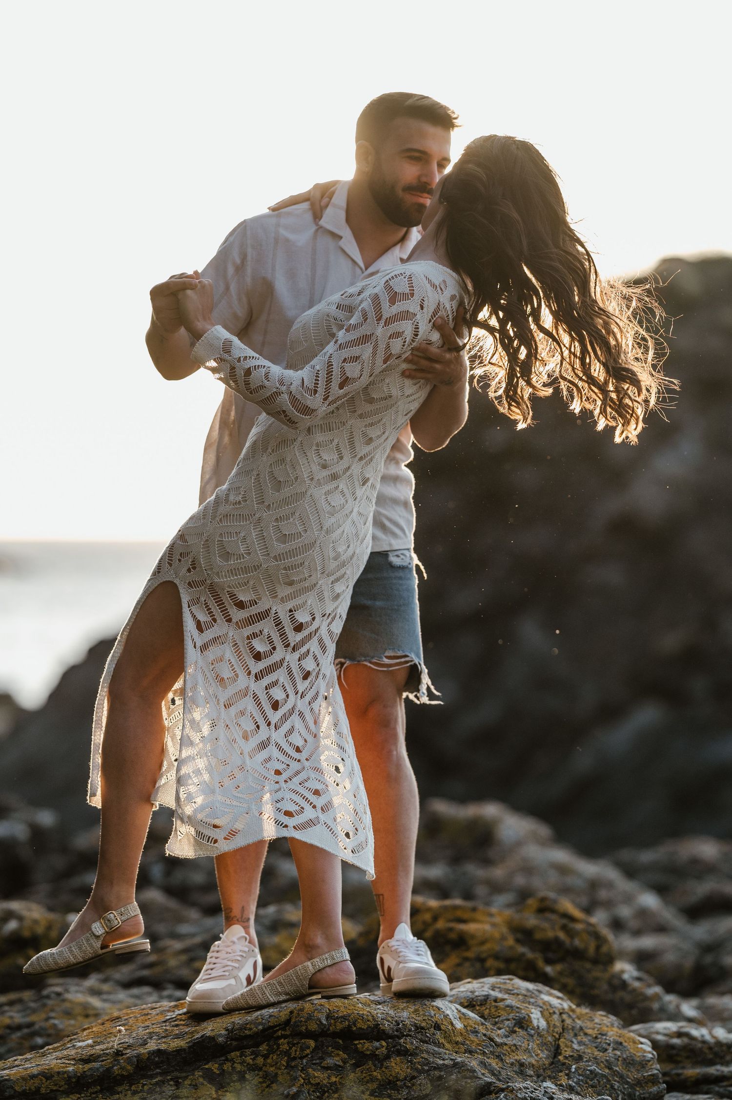 A couple in casual attire share an intimate dance moment among coastal rocks during golden hour.