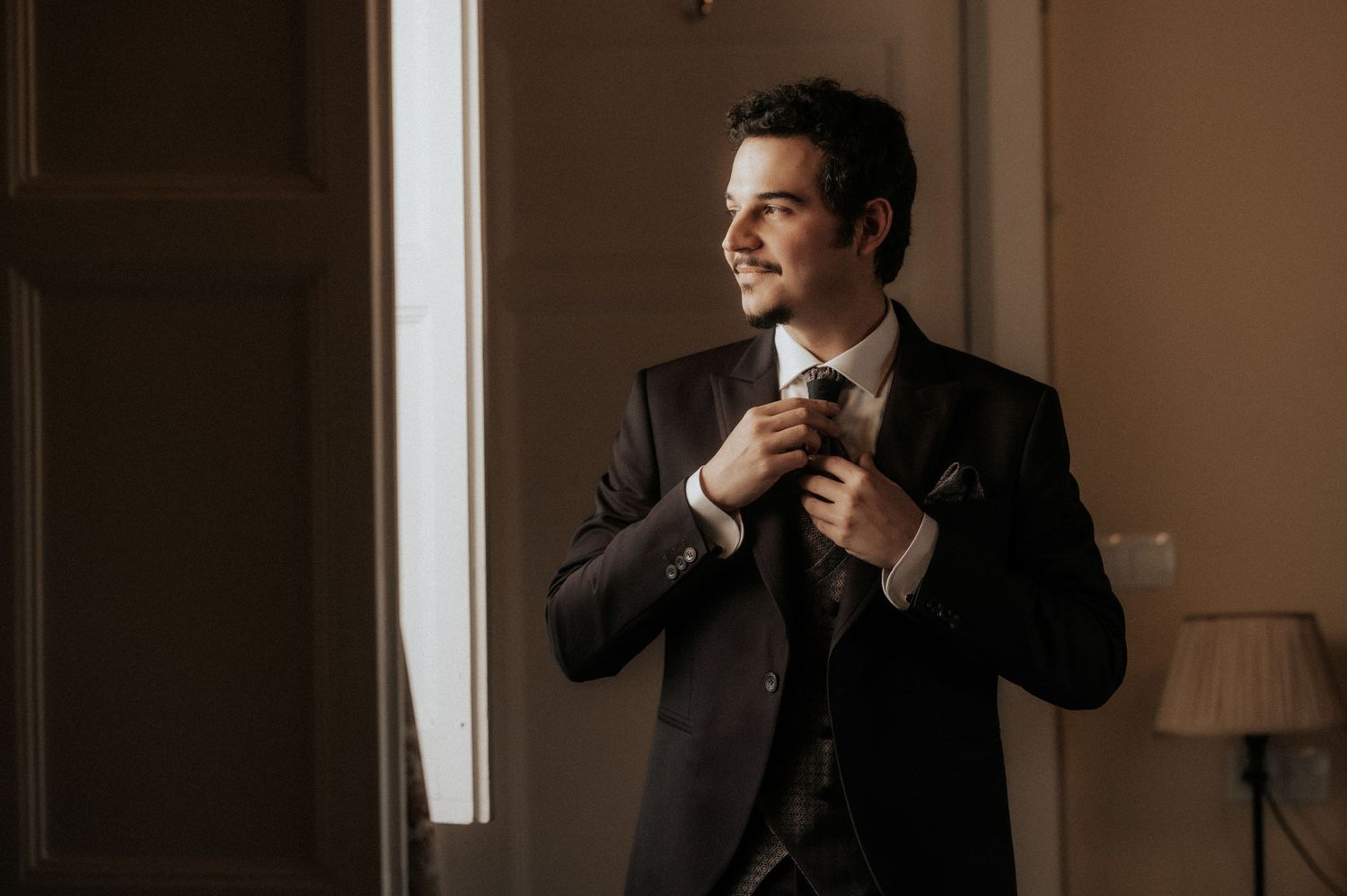 Man in dark suit smiles warmly while standing in a softly lit interior space.
