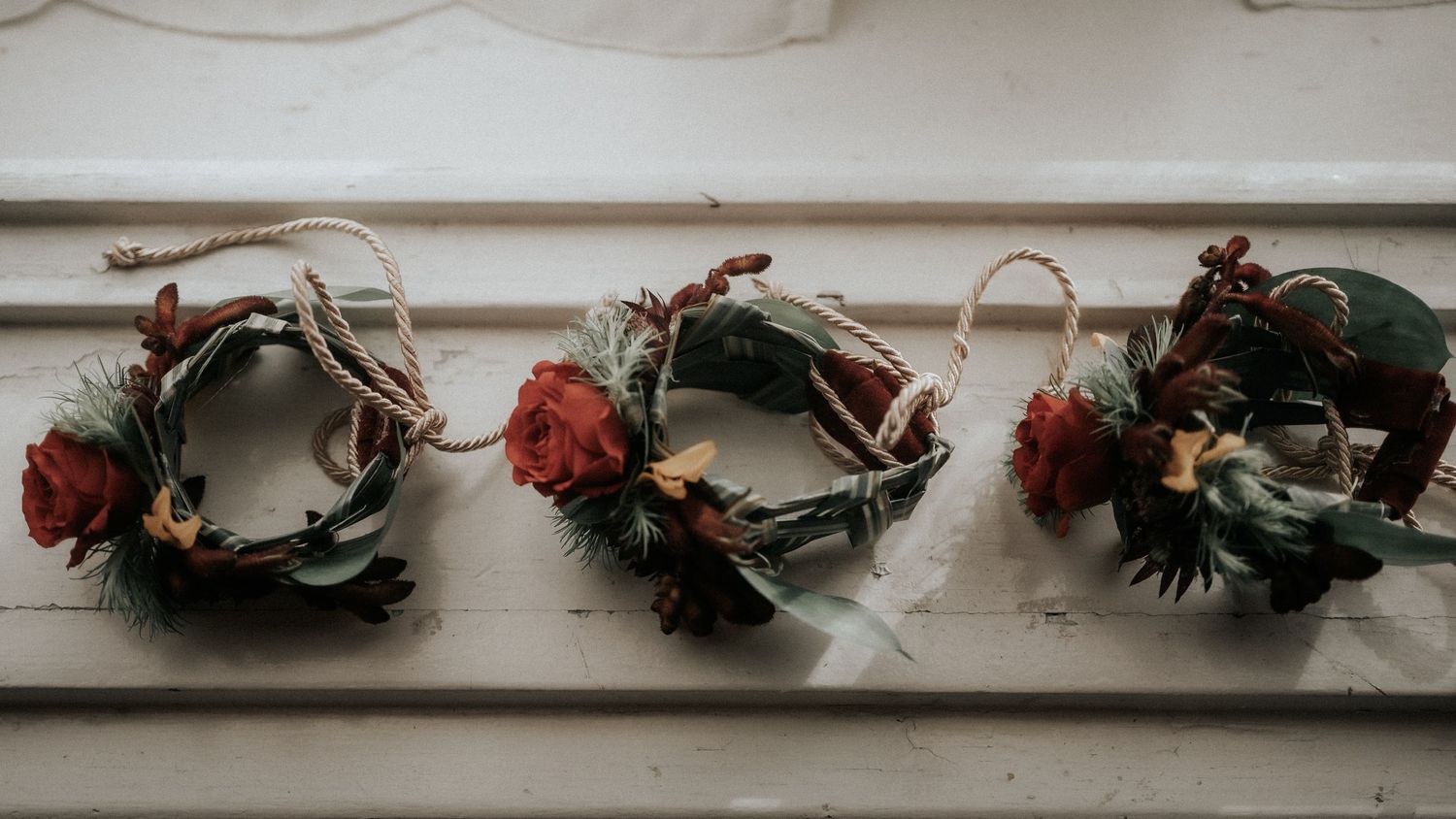 Three rustic wedding boutonnieres with red roses and greenery displayed on a white wooden shelf.