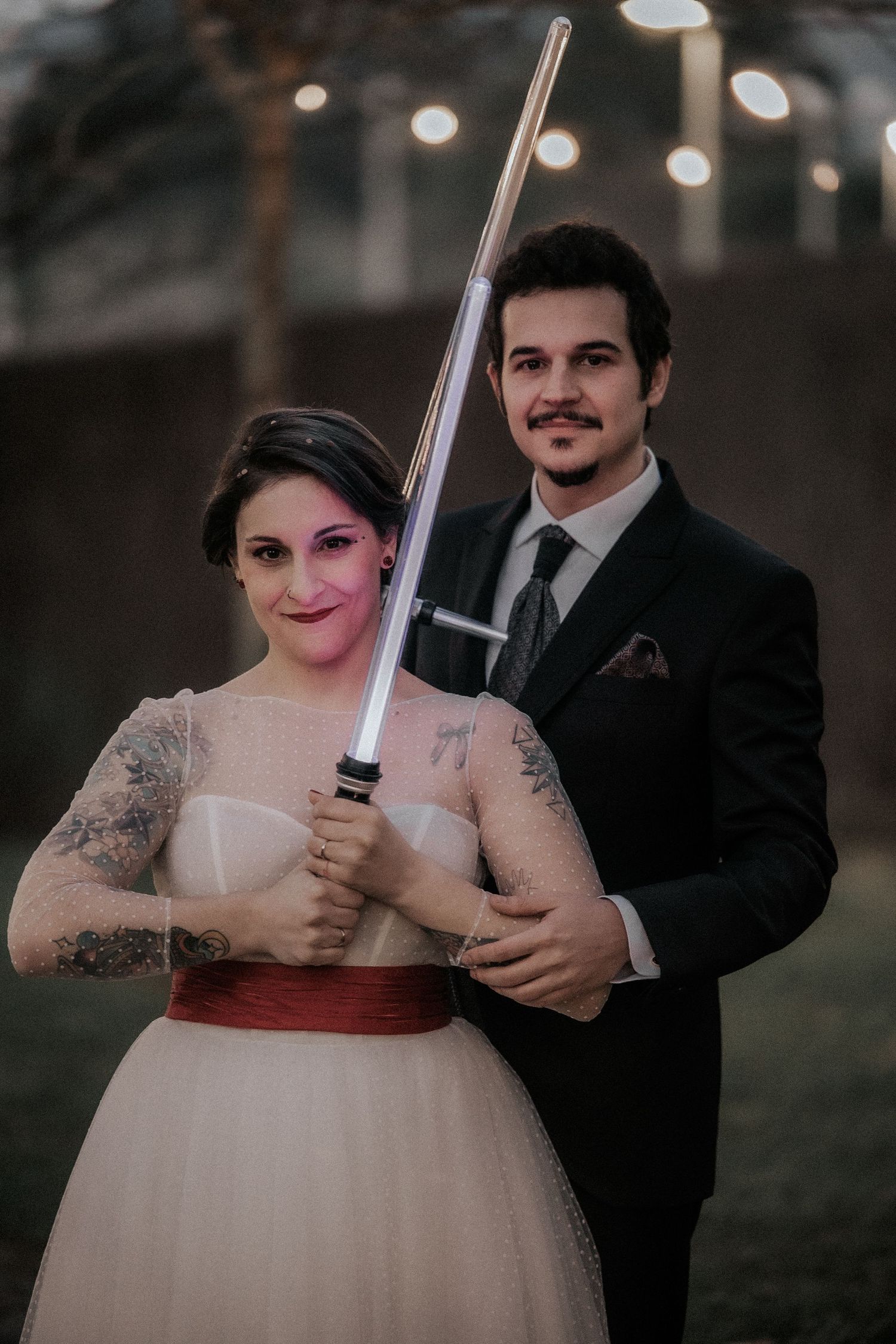 A couple poses at their wedding, with the bride holding a lightsaber while wearing a white dress with red sash.