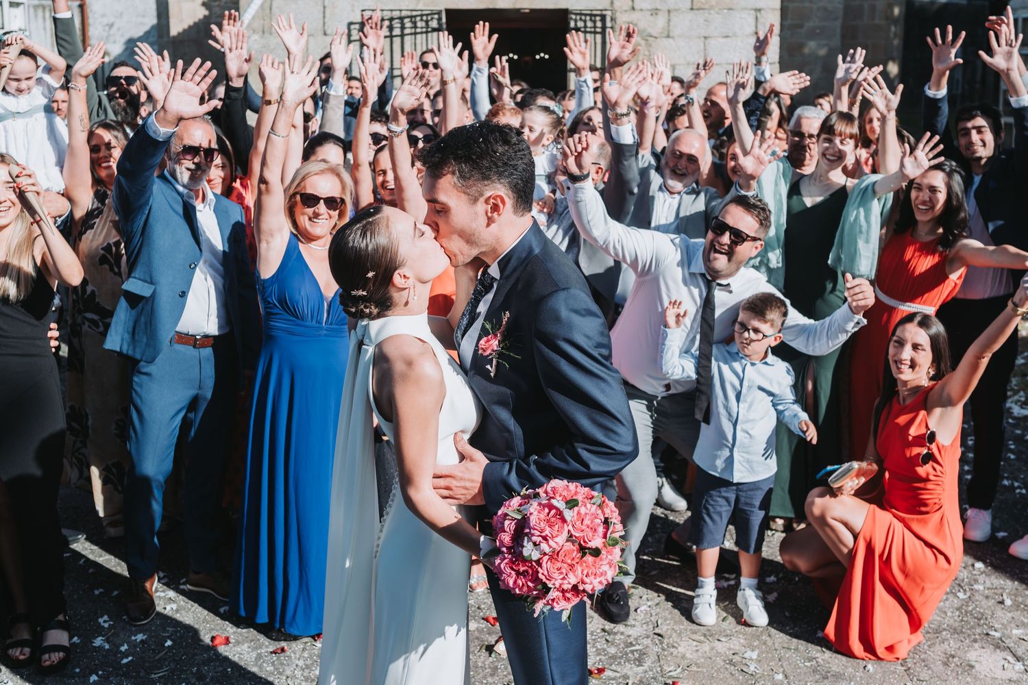 Joyful wedding party and guests raise their hands in celebration outside a stone venue.