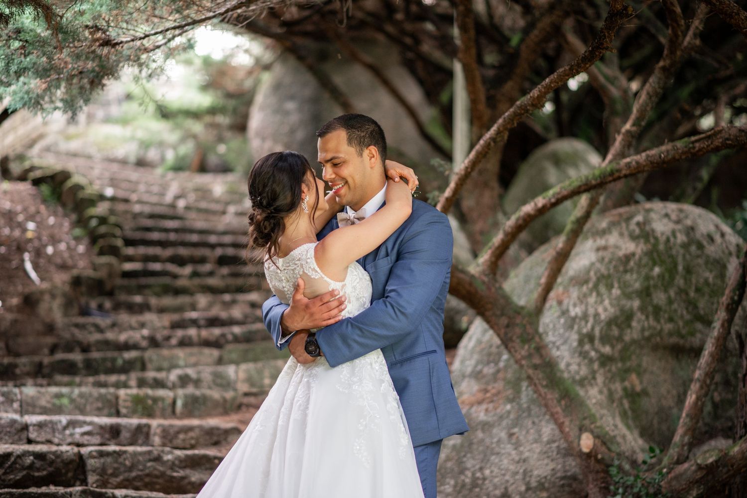 A couple in wedding attire embraces while standing on stone steps surrounded by lush greenery.
