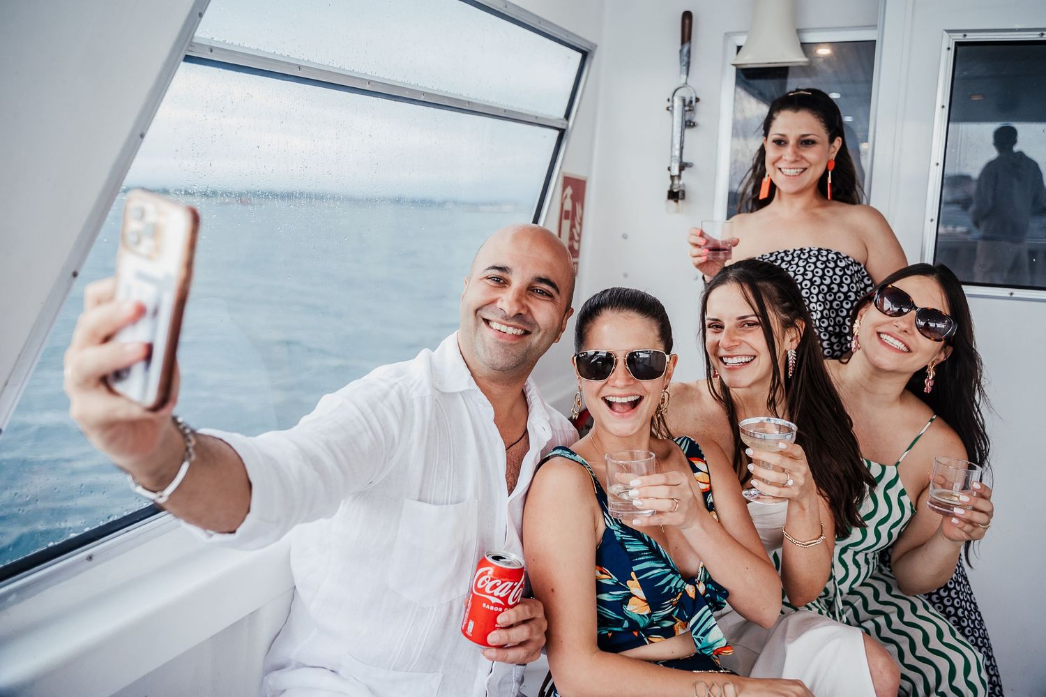 Group of friends taking a selfie while celebrating on a yacht with drinks and smiles.