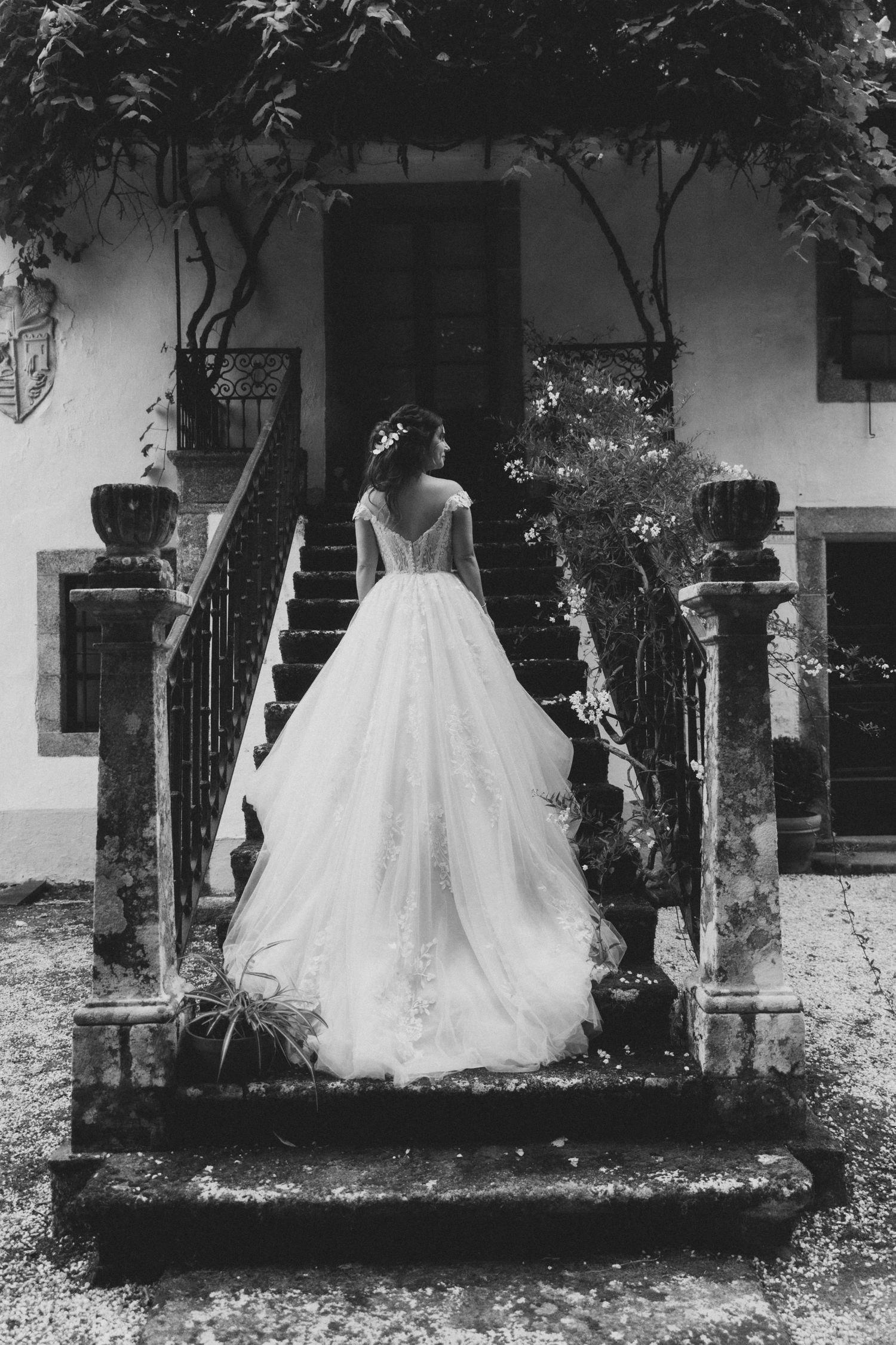 A bride in a flowing white ballgown stands on stone steps in a black and white photograph.