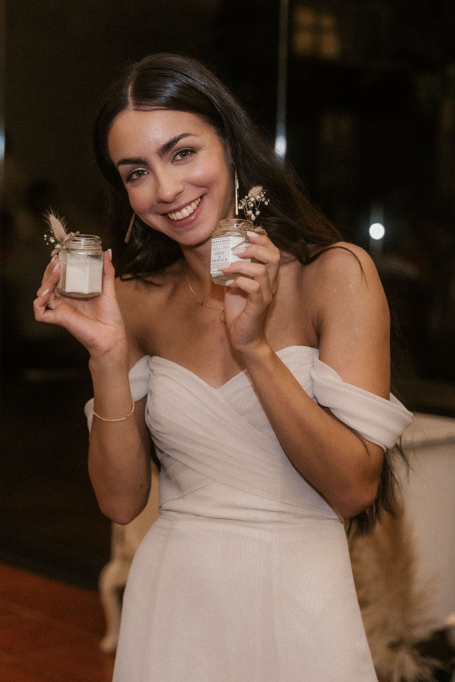 A person in a white off-shoulder dress holds up a small gift box while smiling in a dimly lit room.