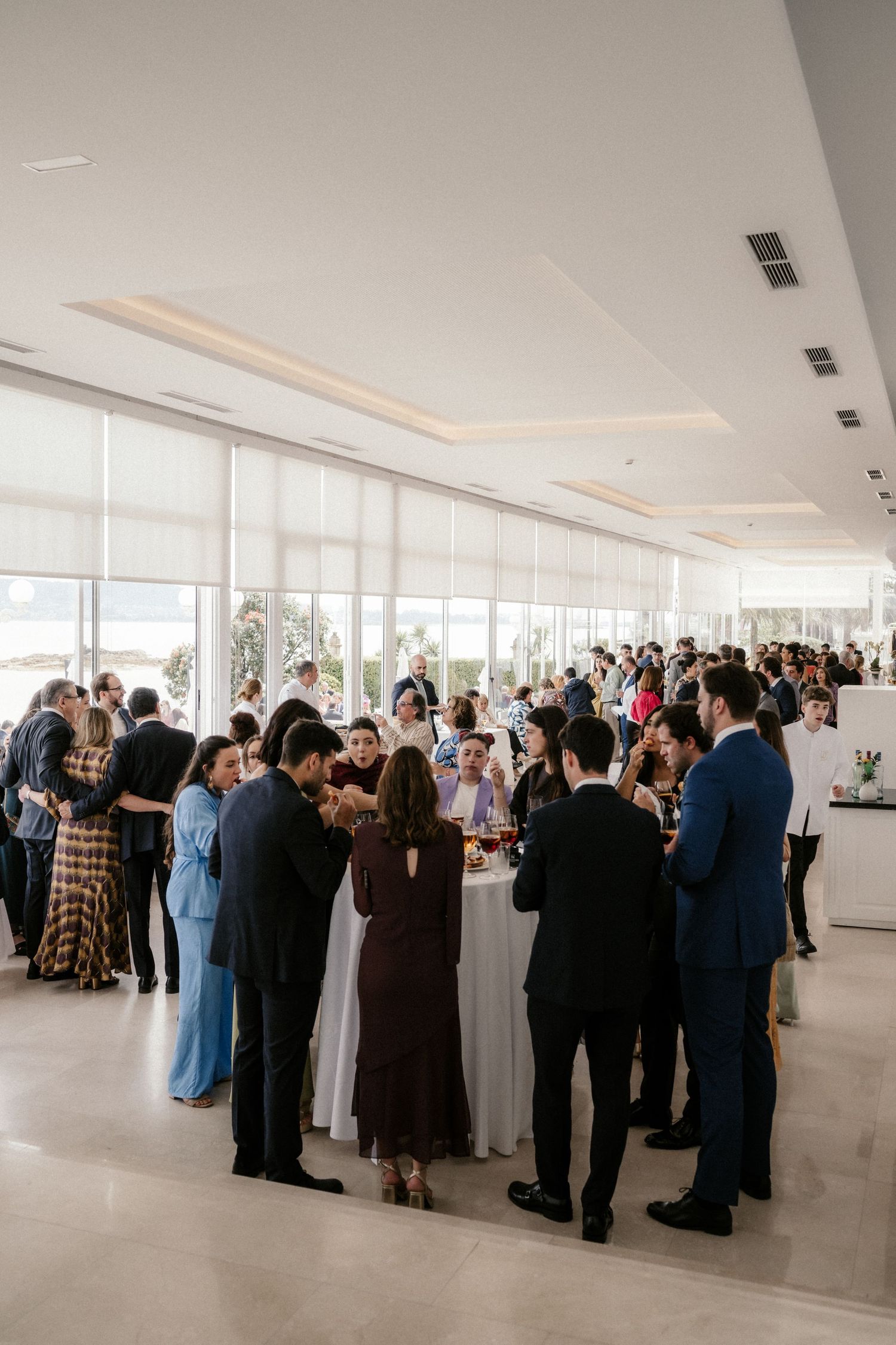 Elegant crowd of guests mingling at a modern event venue with floor-to-ceiling windows and bright interior lighting.