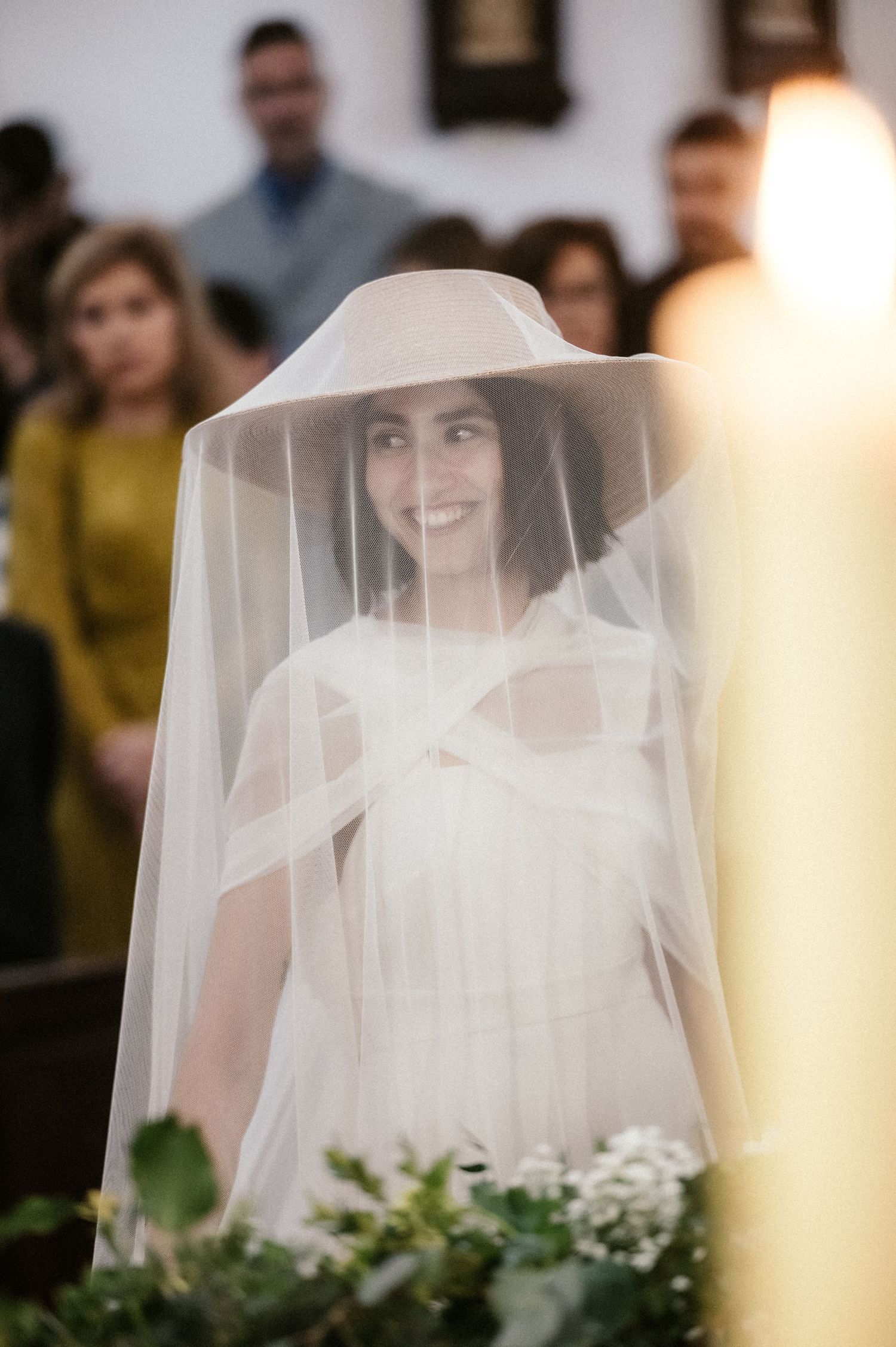 A bride wearing a distinctive wide-brimmed hat veil smiles during her wedding ceremony.