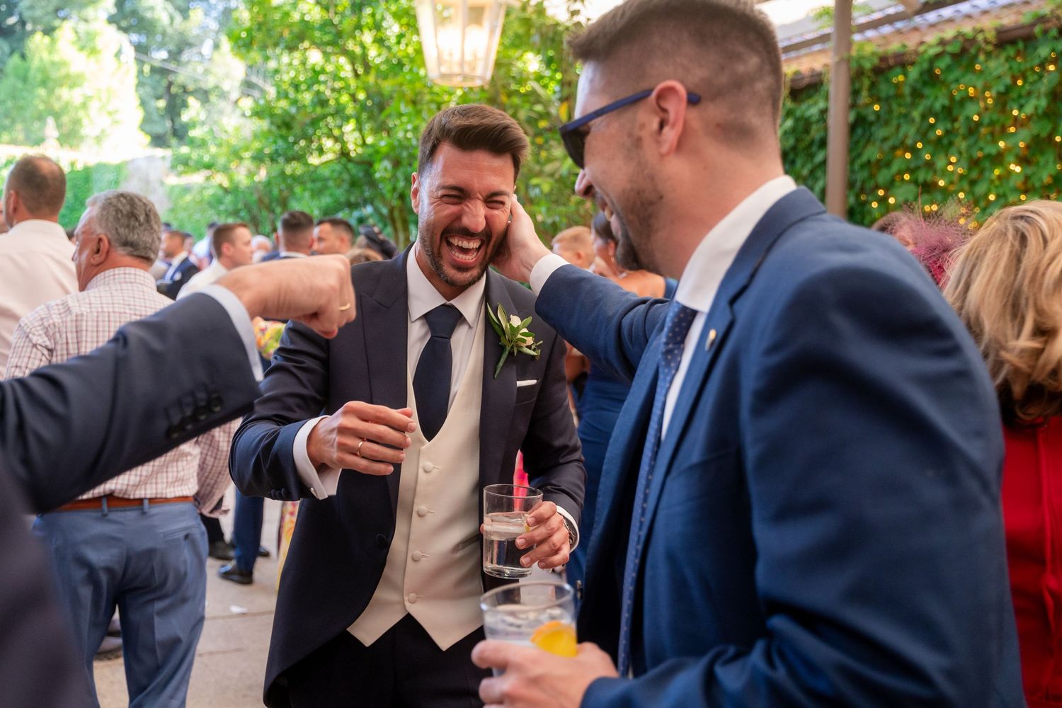 A group of guests in formal attire laugh and socialize at an outdoor event with string lights overhead.