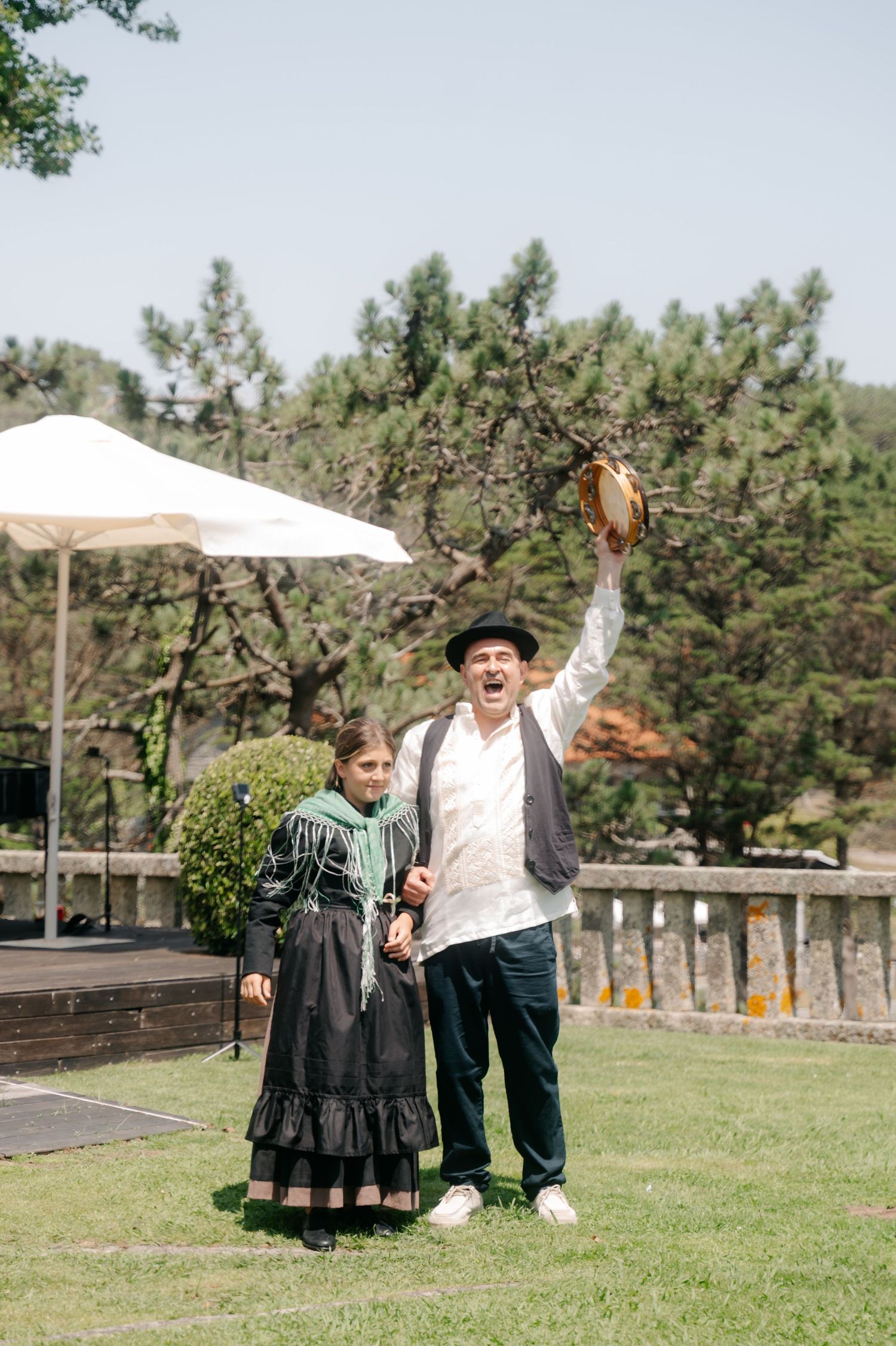 Two people in vintage attire stand on a grassy lawn with pine trees and a white tent in the background.