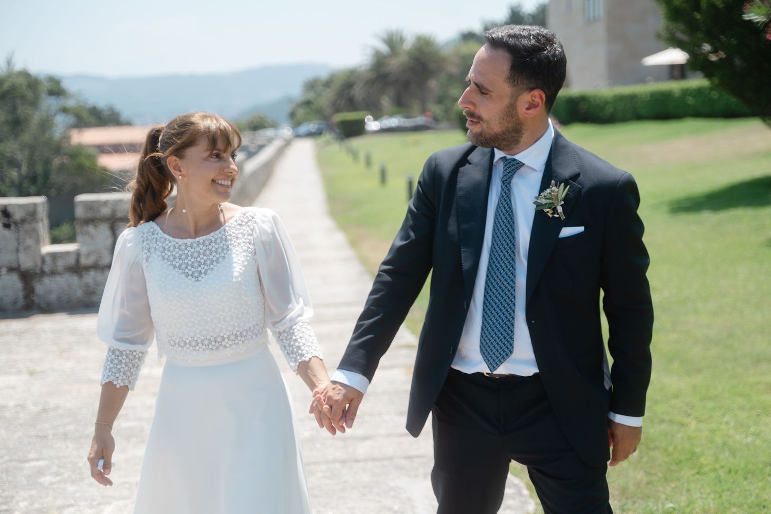 A couple in wedding attire walks together on a grassy path holding hands on their special day.