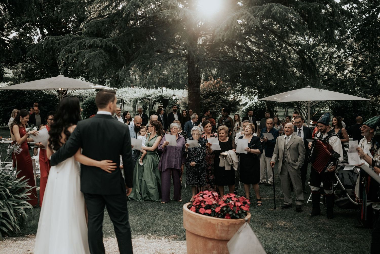 Wedding guests gather outdoors under trees during a backyard garden ceremony at sunset.
