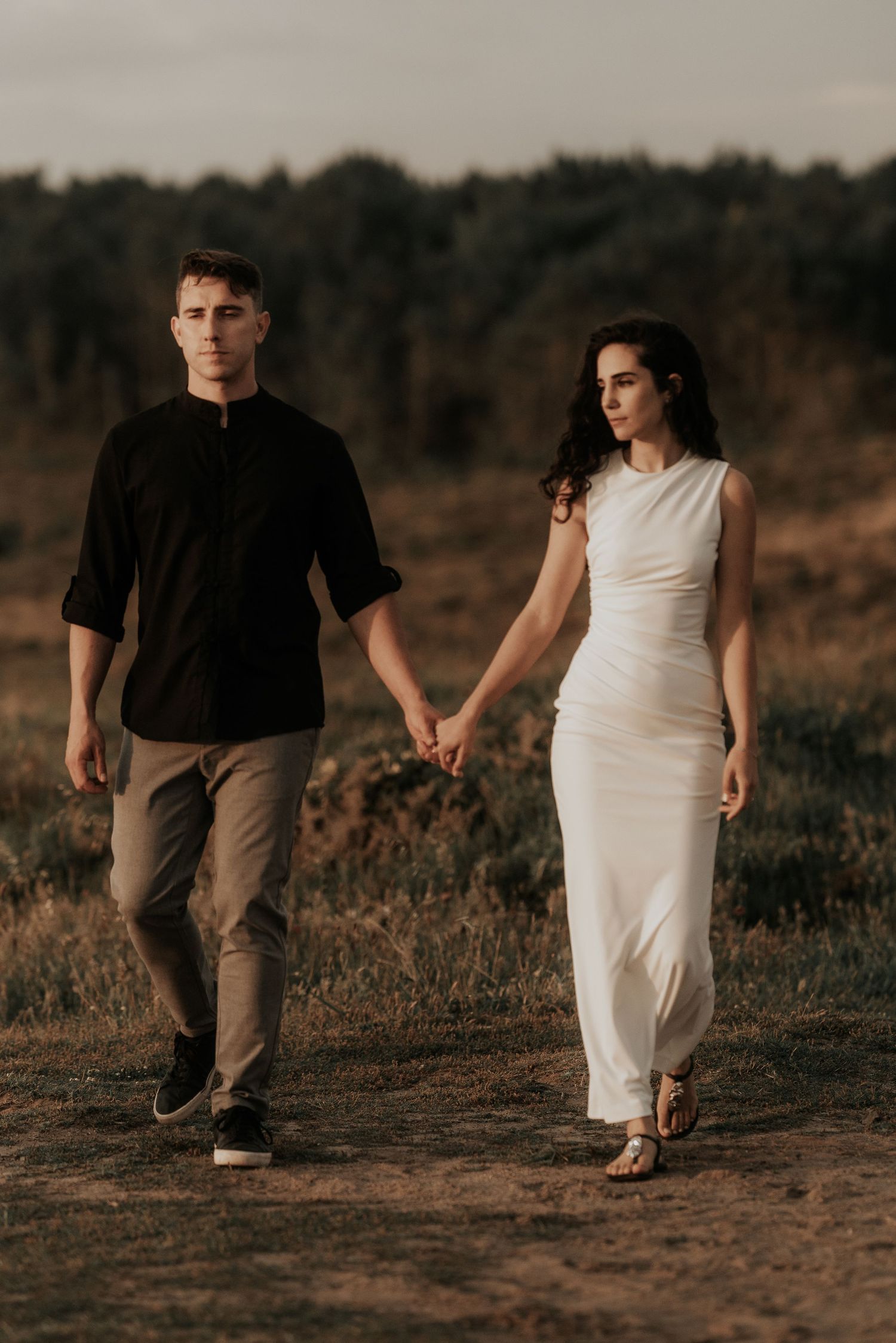 A couple in contrasting black and white outfits walk hand in hand through a rustic outdoor setting at sunset.