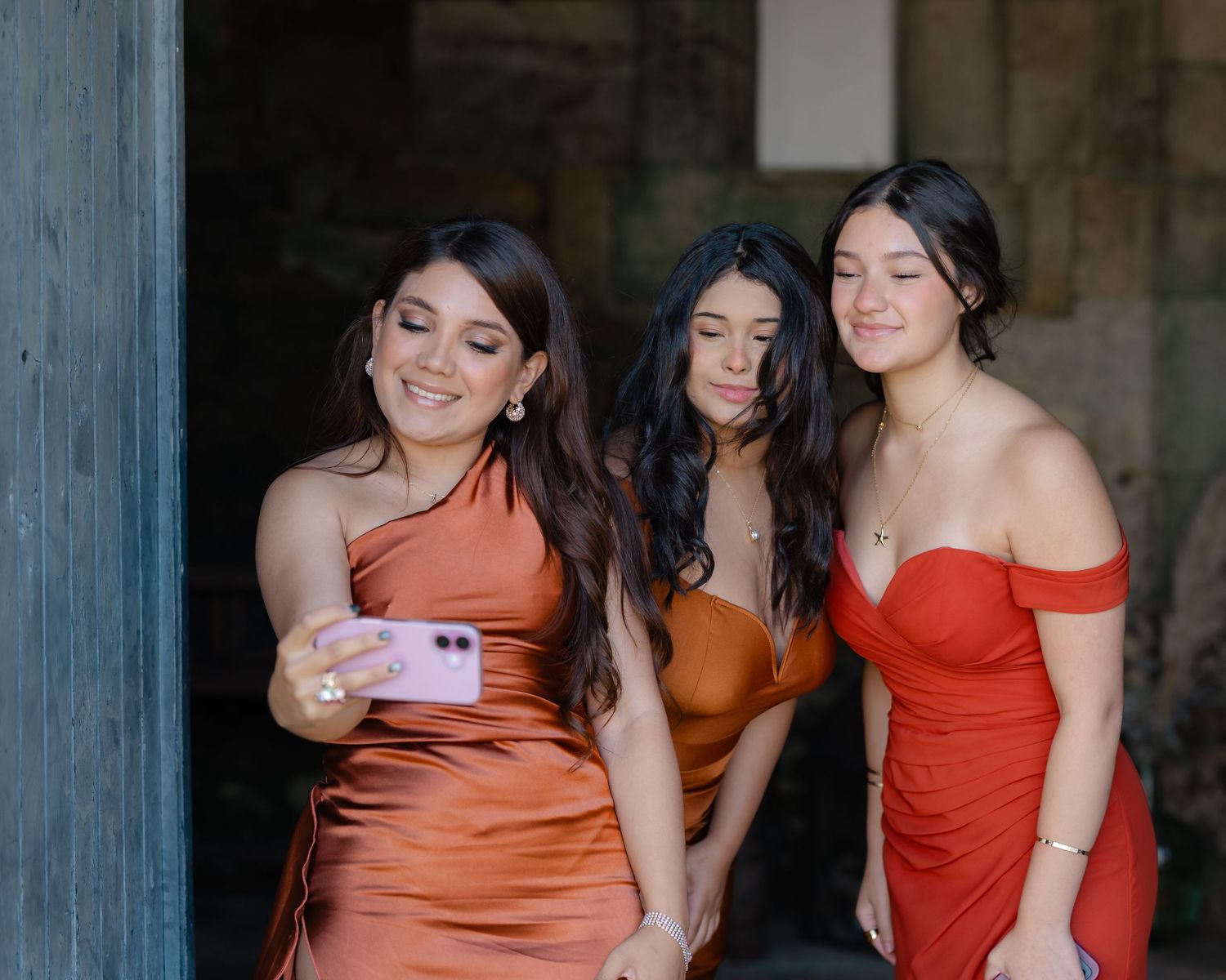 A group of bridesmaids in rust-colored dresses pose together against a rustic blue wall backdrop.