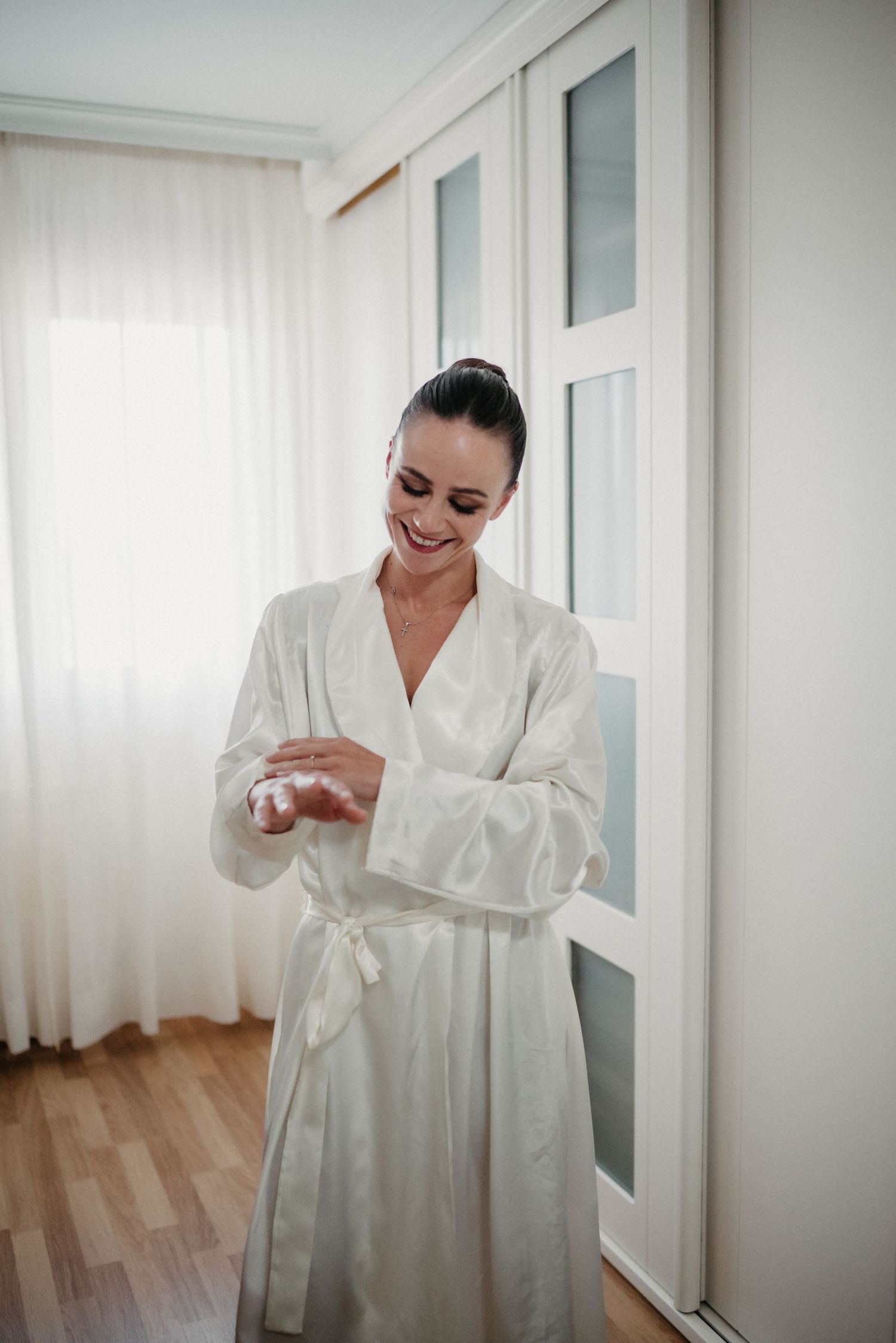 Woman in white bathrobe getting ready in bright modern bedroom with hardwood floors and white decor.