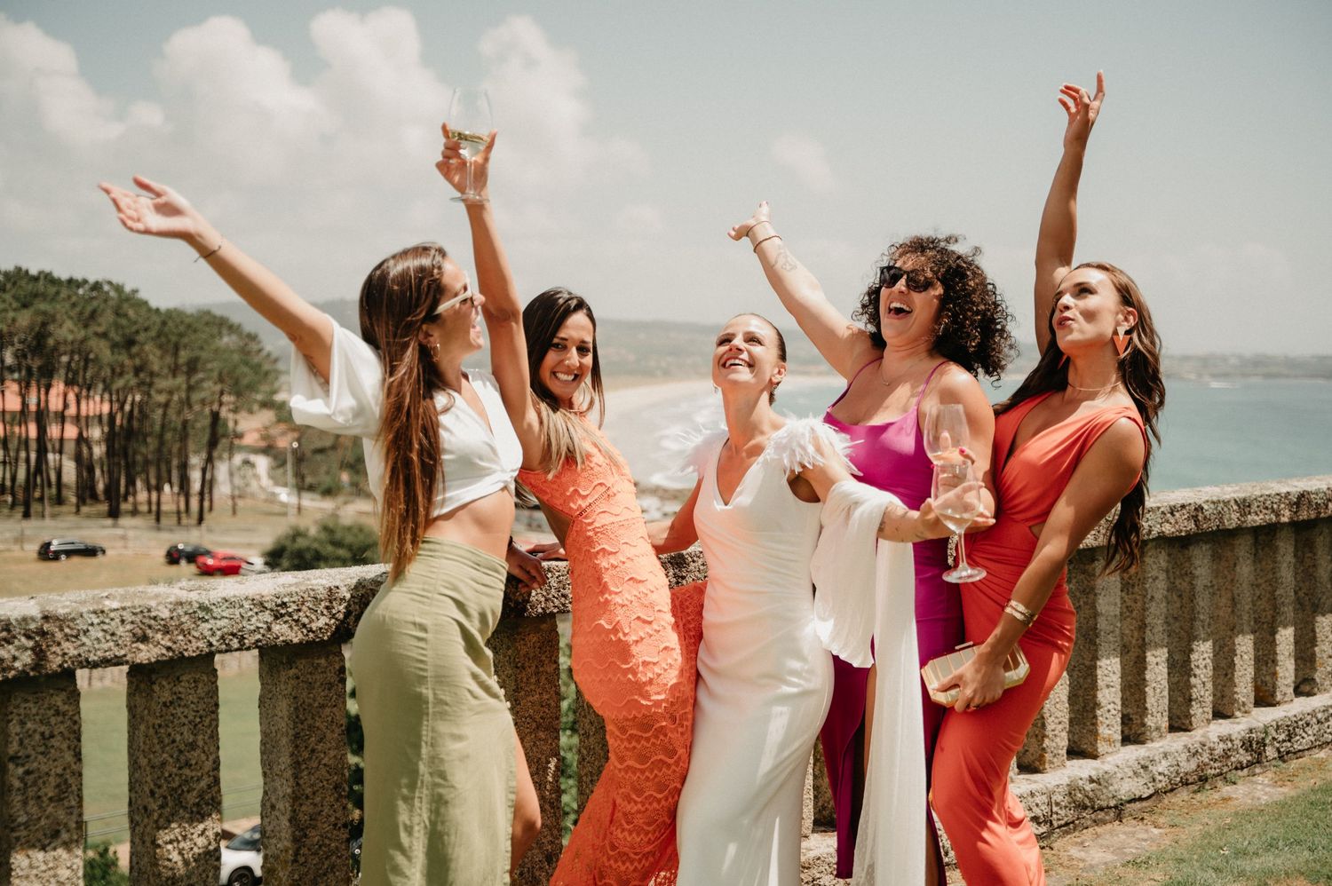Group of bridesmaids in colorful dresses raise their arms joyfully on a sunny beach overlook.