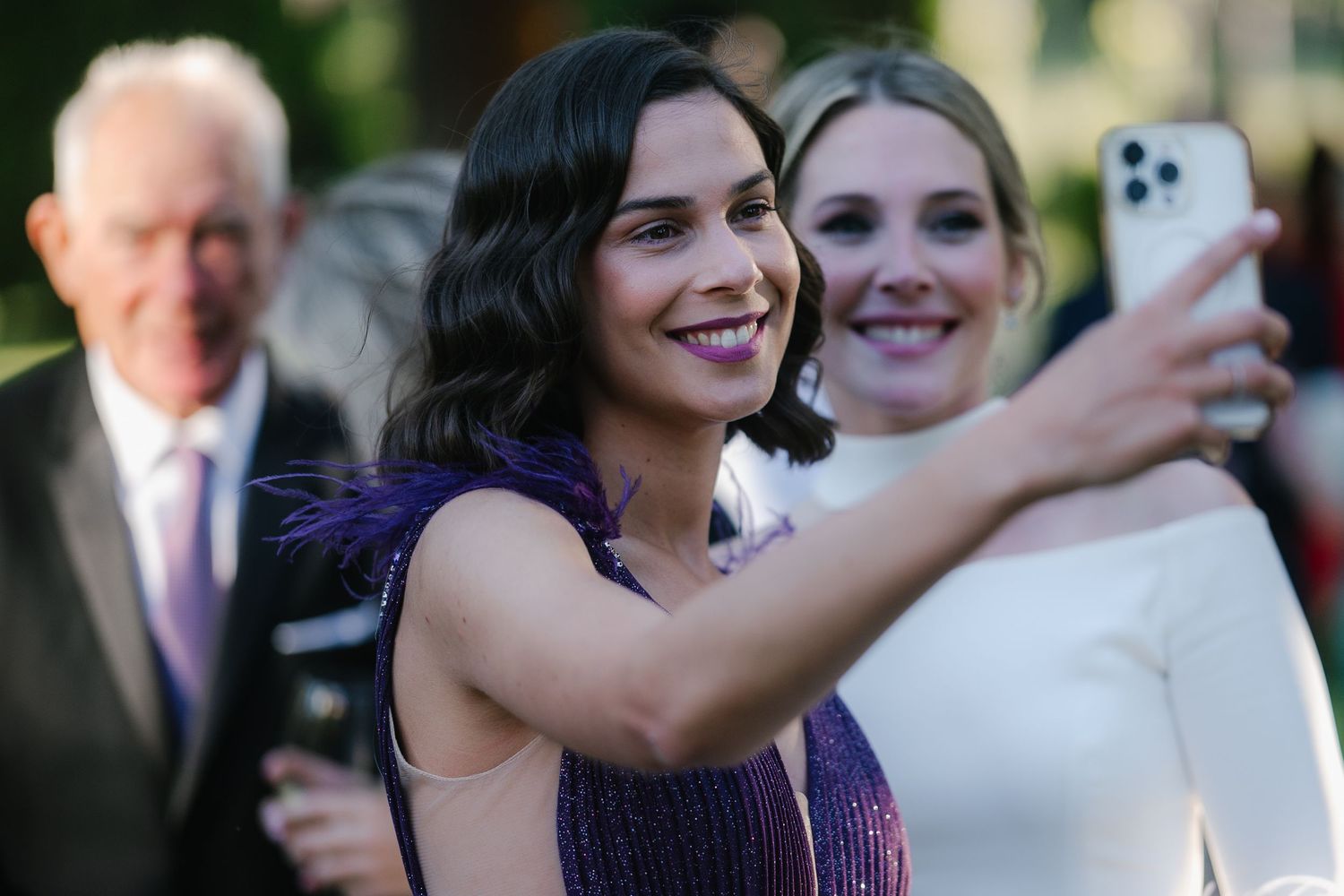 Wedding guests gather outdoors for group selfies and photos during a celebration in a garden setting.
