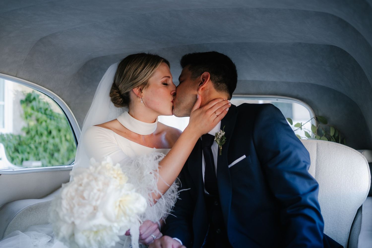 A couple shares a romantic kiss in a vintage car on their wedding day, with the bride in white and groom in navy blue.