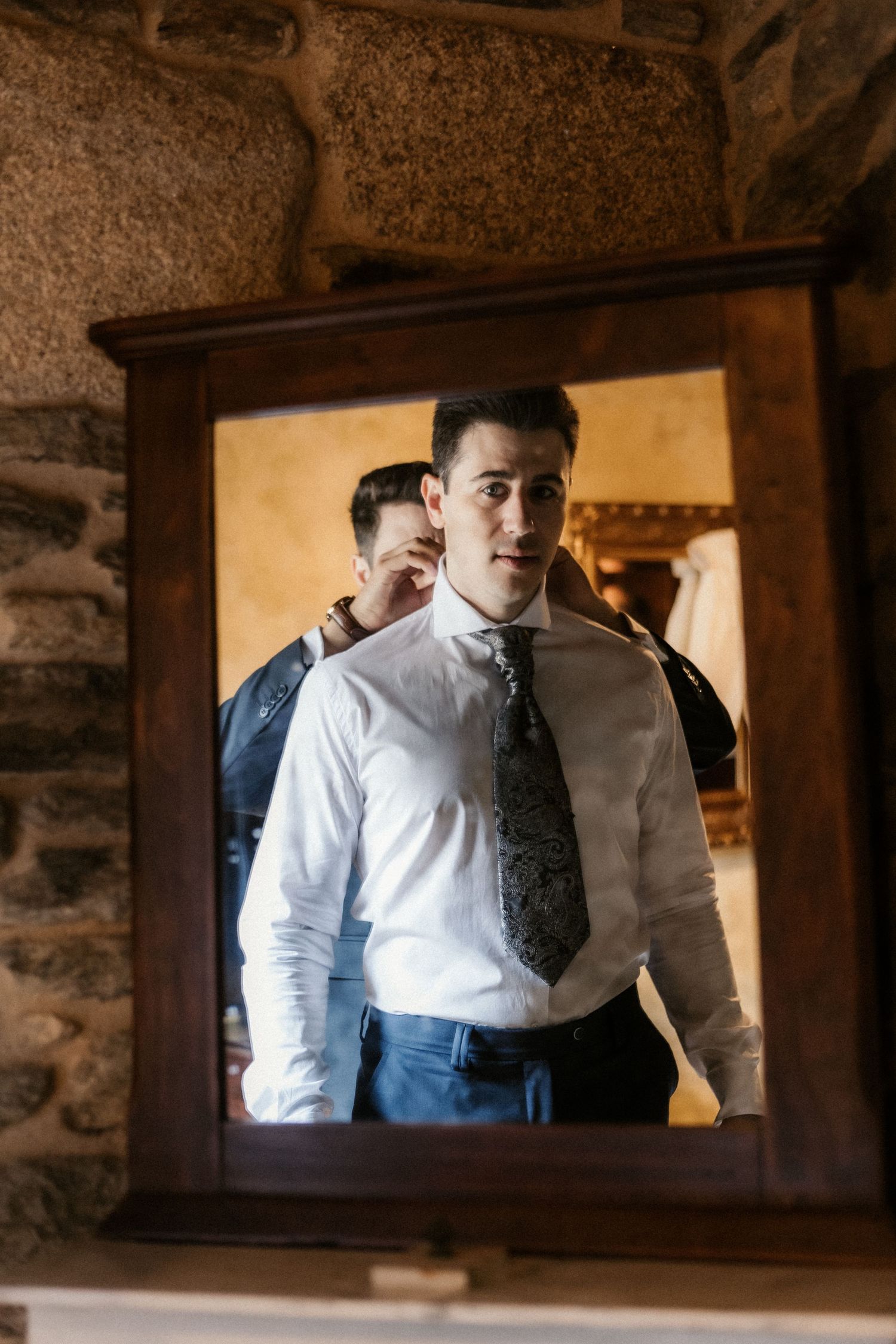 A series of mirror reflections show someone in formal attire with a tie against a rustic stone wall background.