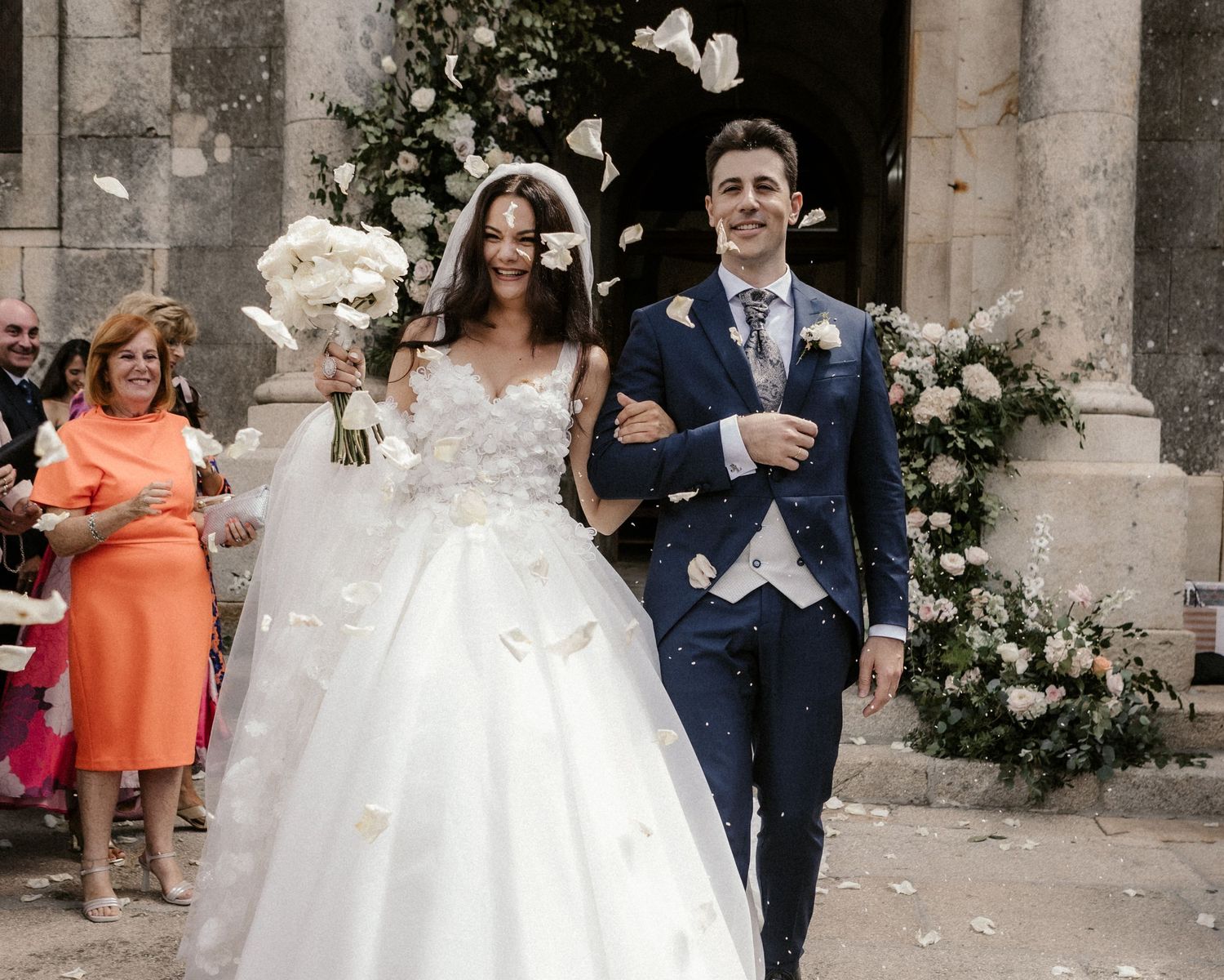 Wedding party and guests celebrate with confetti outside historic stone church entrance.
