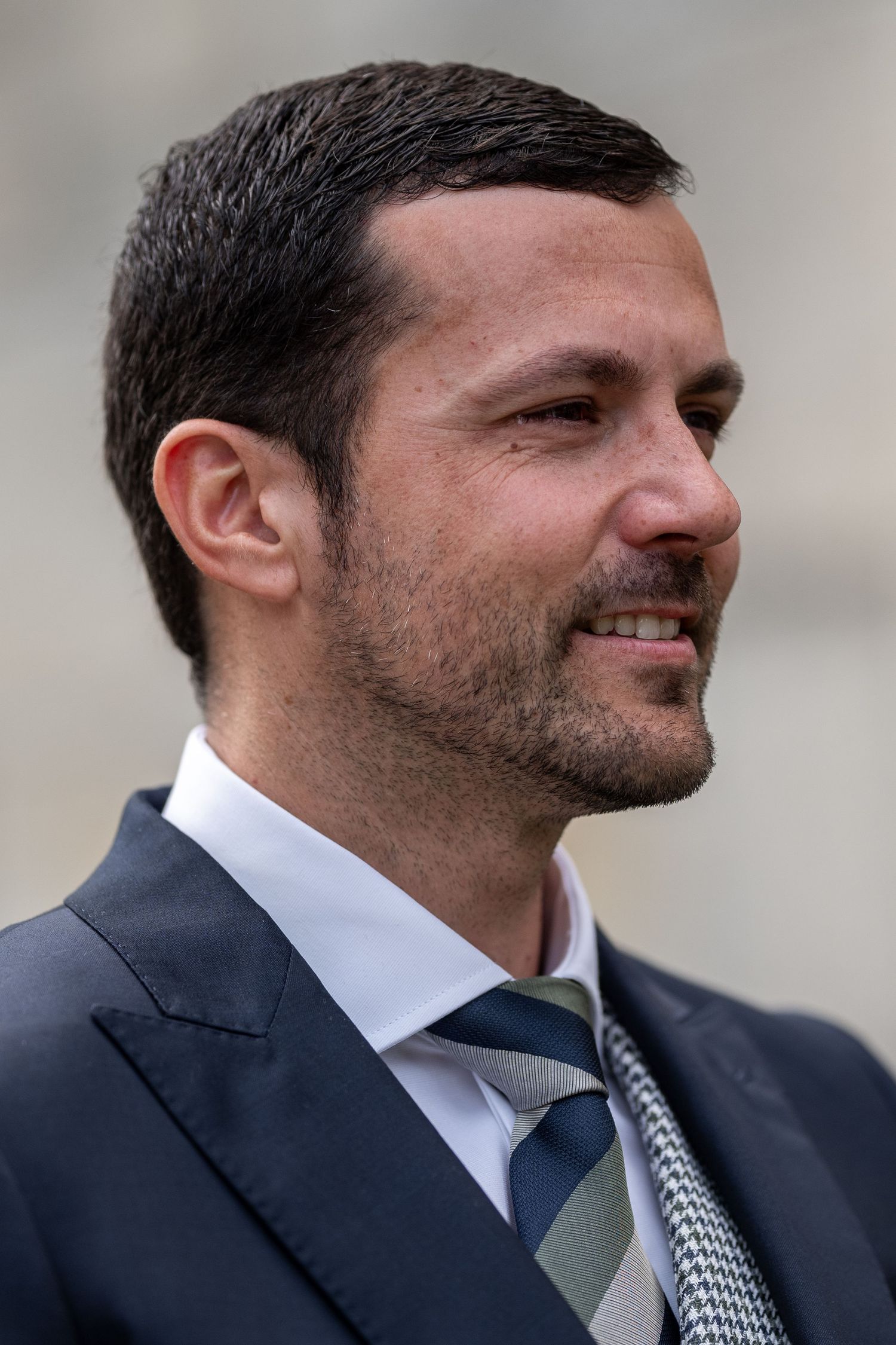 Side profile view of a groom in a navy suit with striped tie smiling during ceremony.