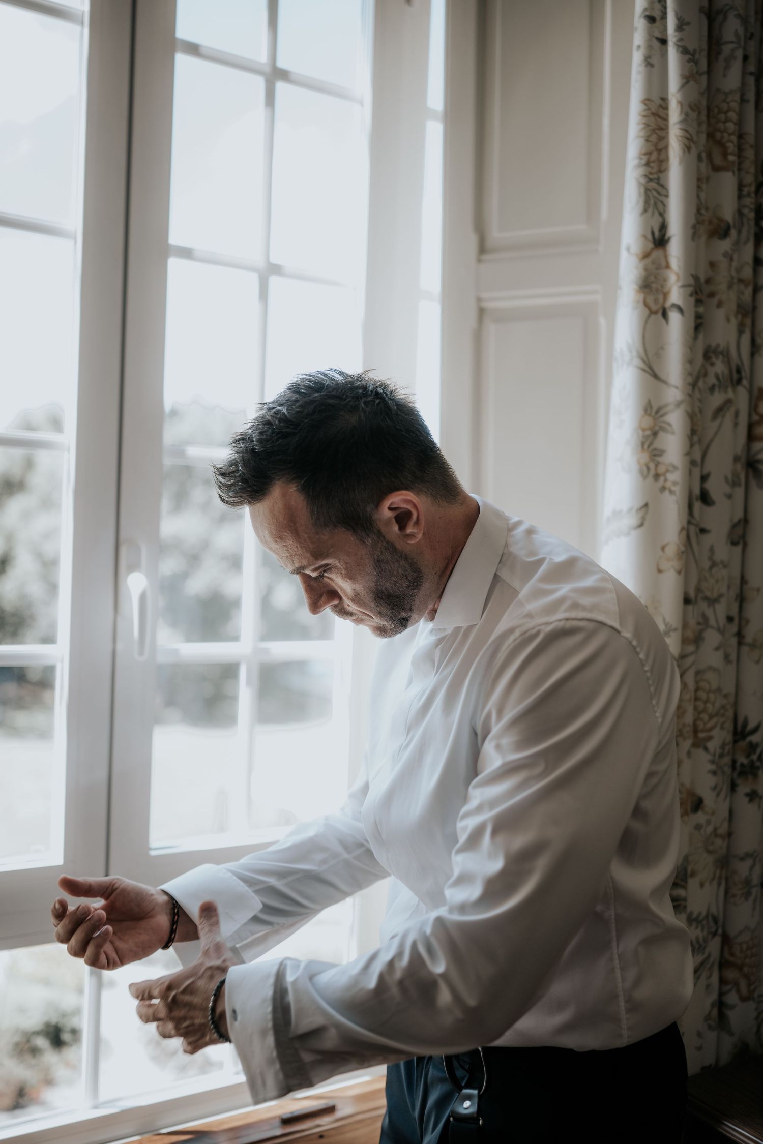 A person in a white dress shirt adjusts their cuffs while standing by a bright window with floral curtains.