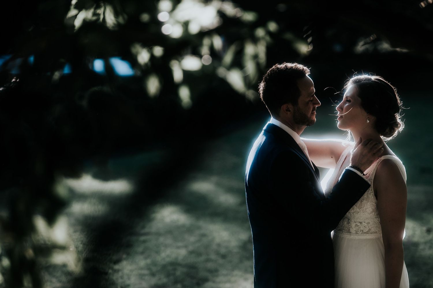 A couple shares an intimate moment in a garden setting with dappled sunlight filtering through trees.