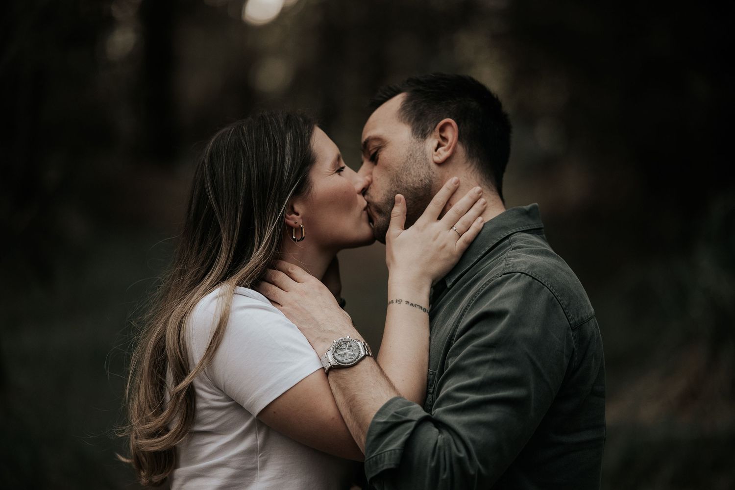 A romantic couple shares an intimate kiss in a moody outdoor setting with dark bokeh lighting effects in the background.