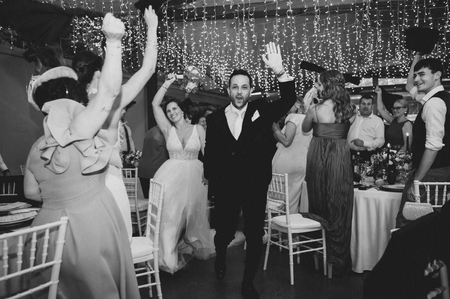 Wedding guests dance and celebrate under twinkling lights at an elegant indoor reception venue.