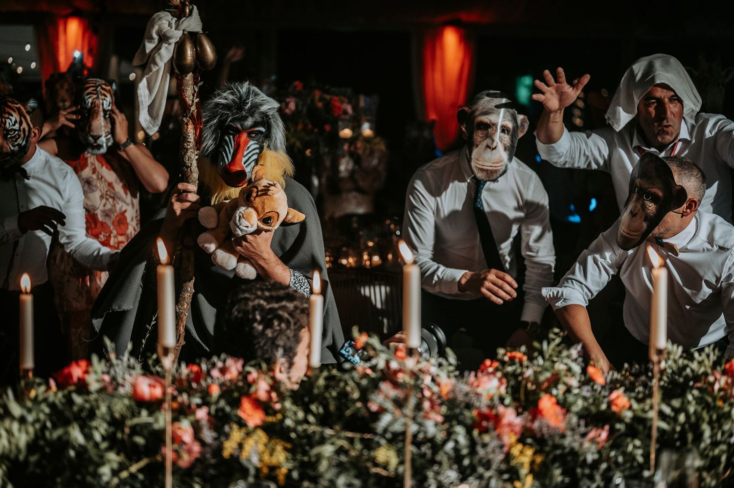 Wedding guests react emotionally during reception behind floral decorations with red lanterns glowing in background.