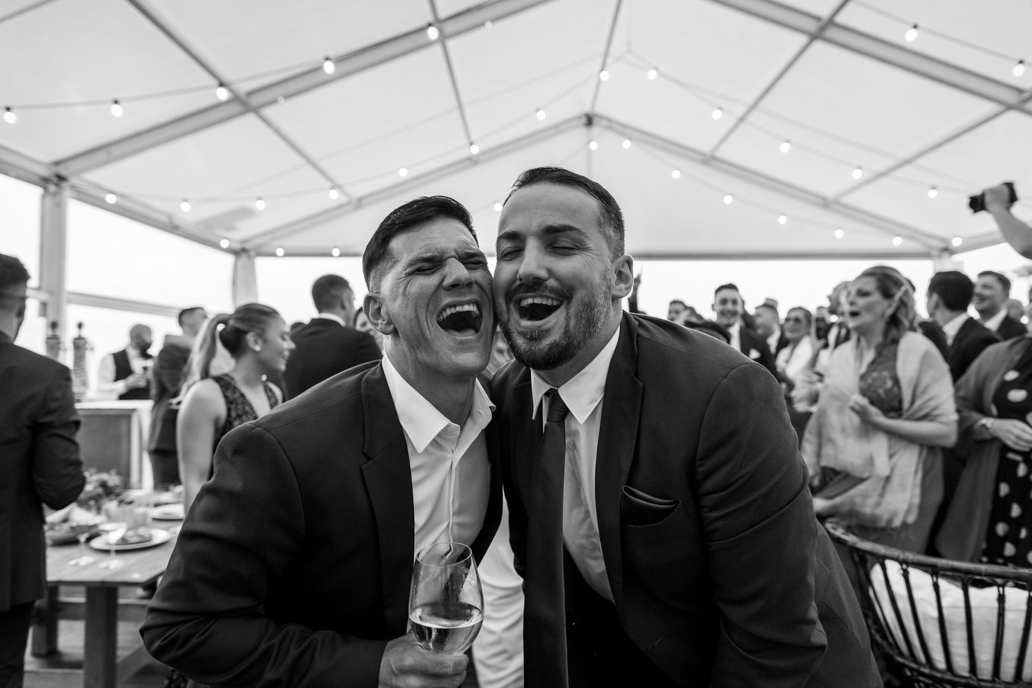 Black and white wedding reception photo showing guests laughing and enjoying themselves at tables under a tent.
