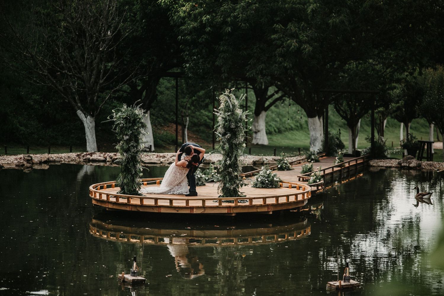 Wedding couple stands beside a serene pond with ornate stone fountain in a peaceful garden landscape.