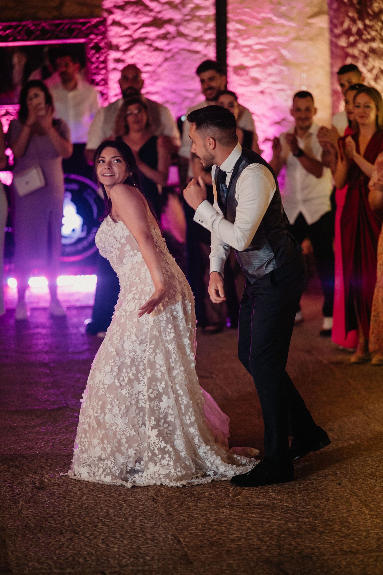 A couple dances together at their wedding reception under pink and purple mood lighting on a stone floor.