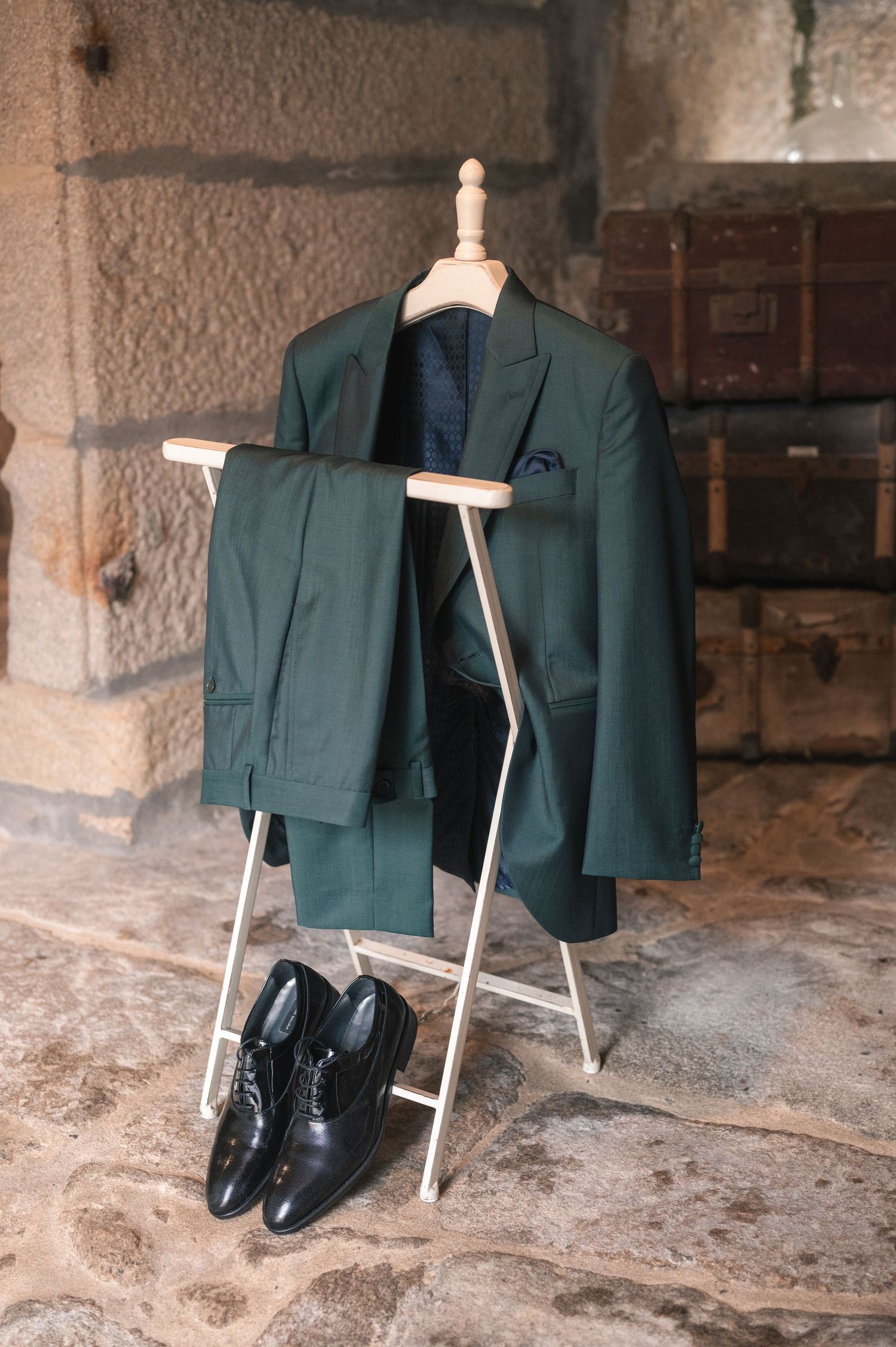 A green formal suit and black dress shoes displayed on a stone floor with an ironing board.