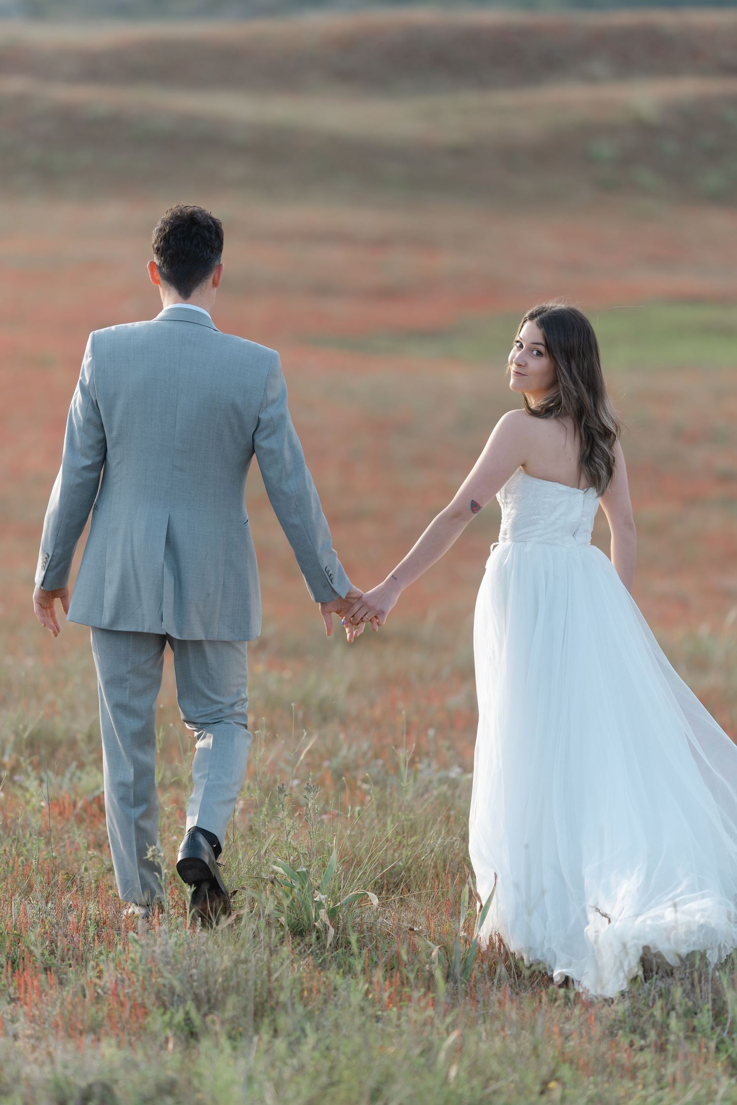 Bride and groom holding hands walking through a red wildflower field, bride in white strapless gown, groom in gray suit.