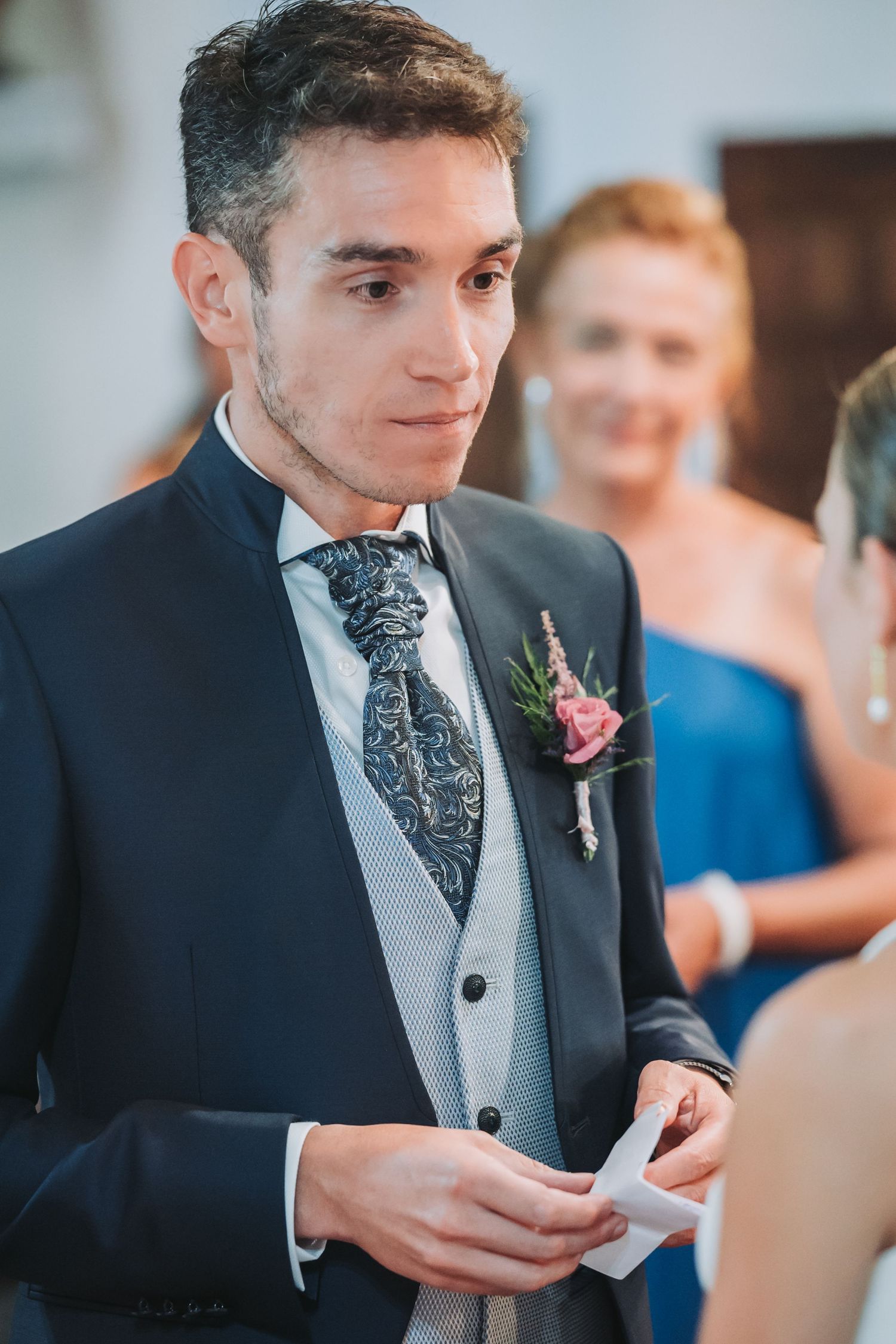 Groom in dark suit and patterned tie reads a heartfelt note during wedding ceremony.