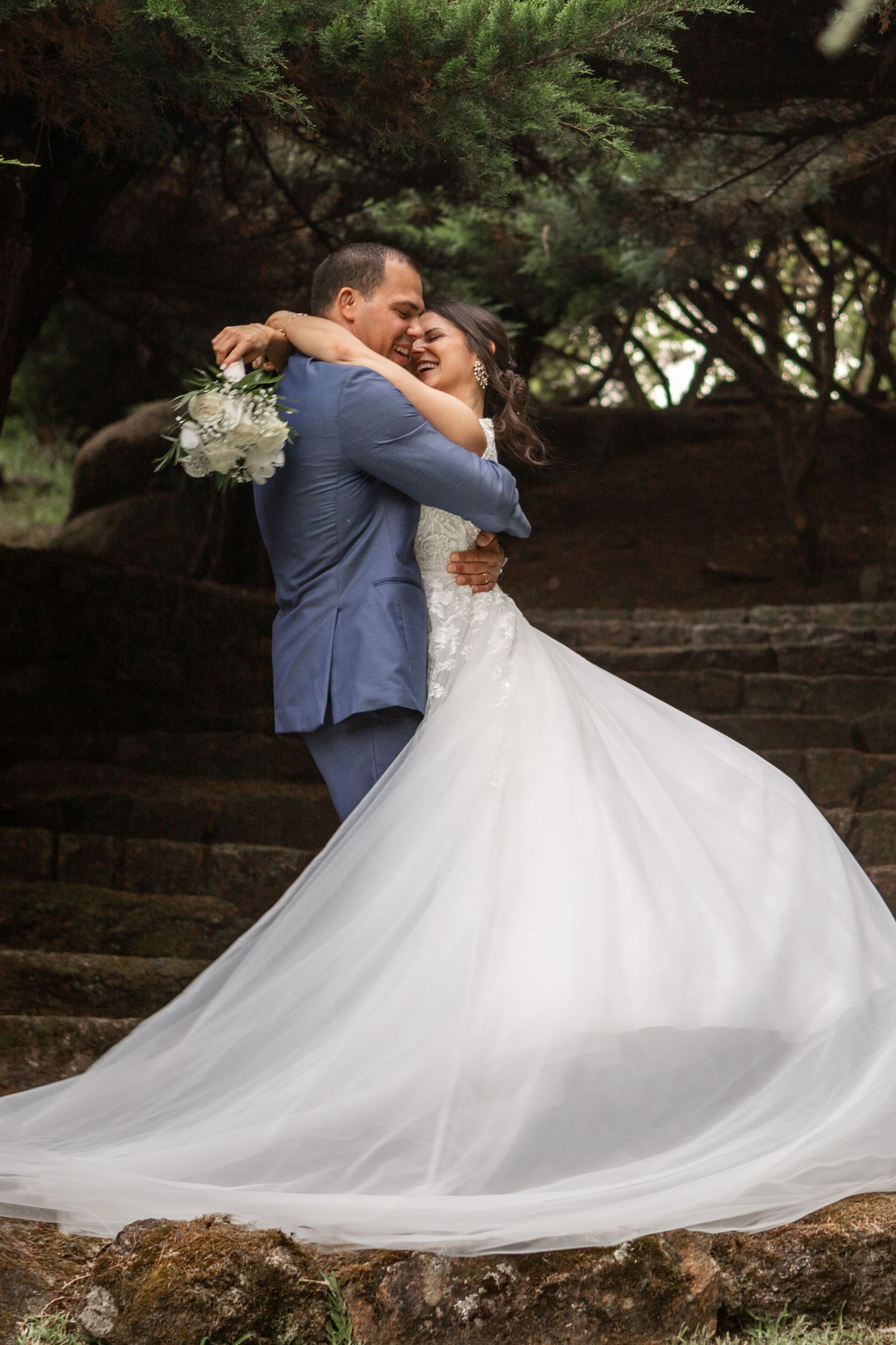 A couple in wedding attire dances and spins together on stone steps surrounded by lush greenery.