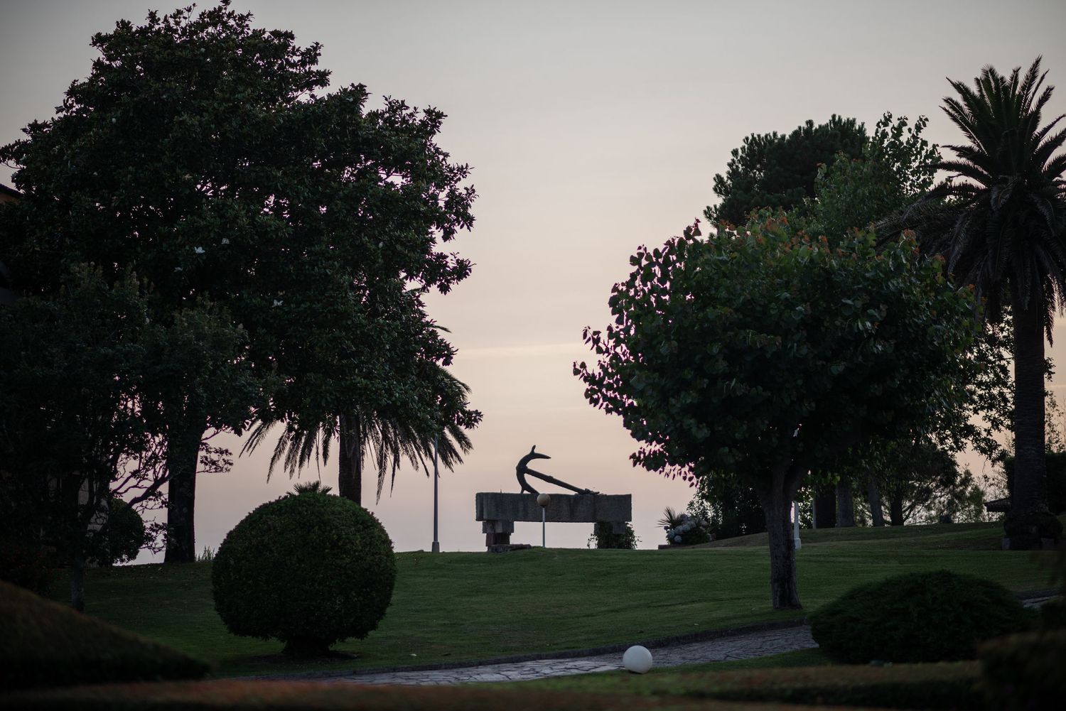 A scenic coastal path with palm trees and lush greenery against a dusky sky at sunset.