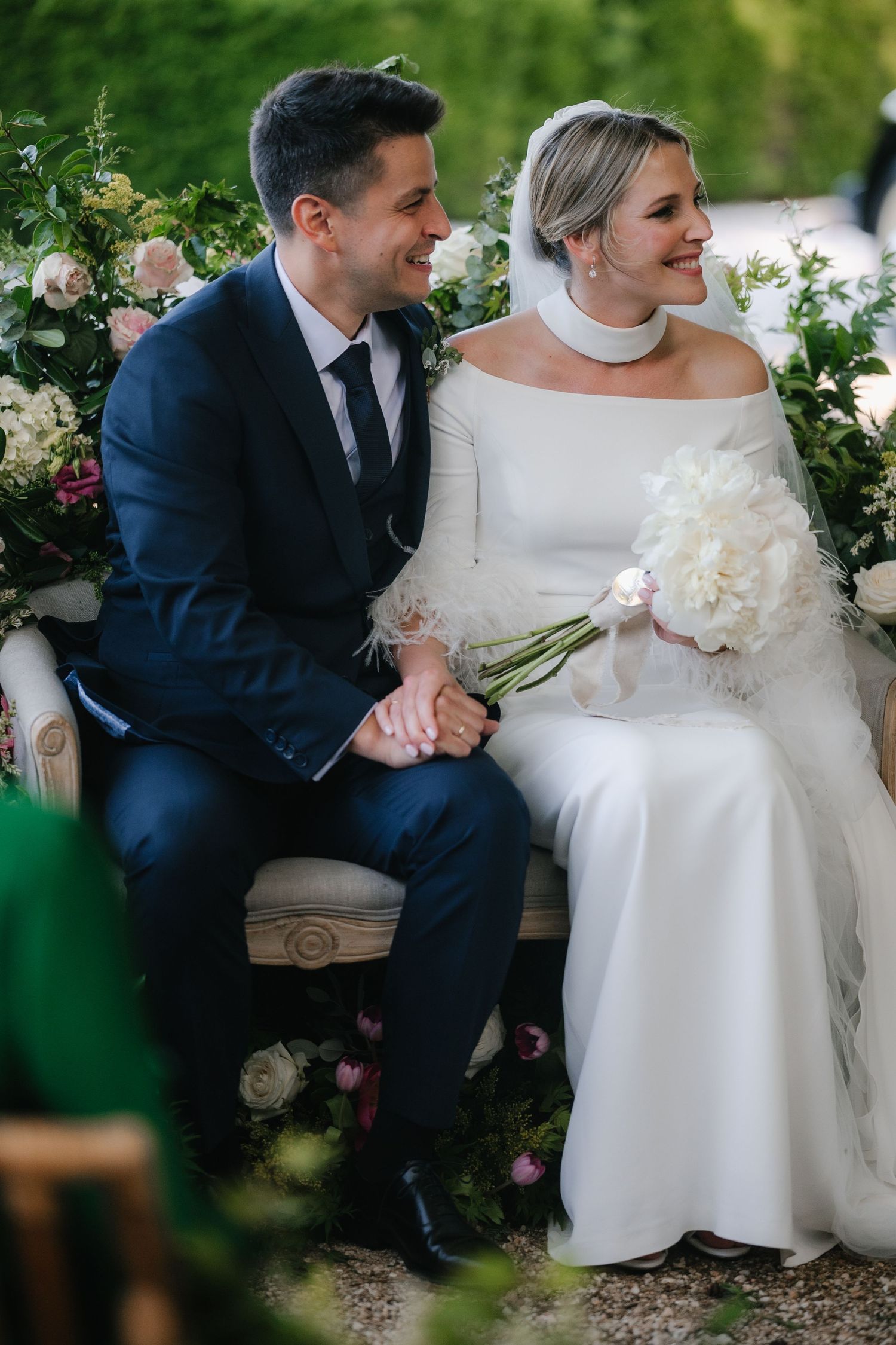 A joyful couple shares a candid moment on their wedding day surrounded by floral arrangements in an outdoor garden setting.