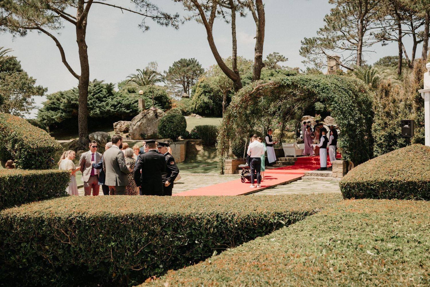 A scenic outdoor garden wedding ceremony setup with red carpet aisle surrounded by manicured hedges and trees.