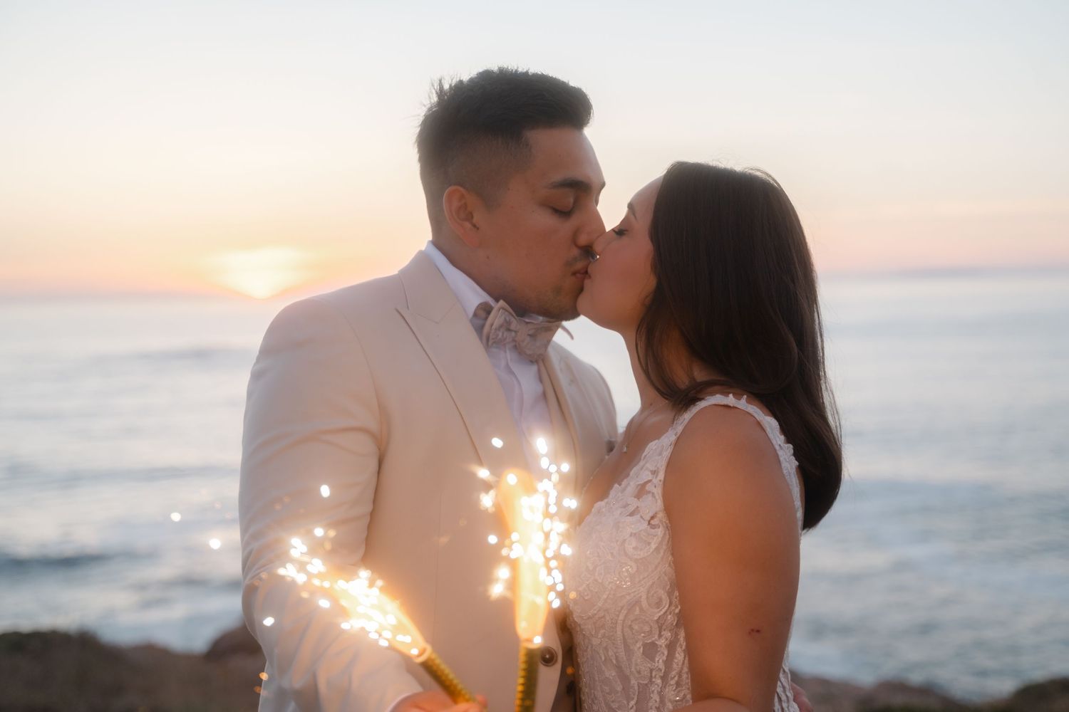 Newlyweds share intimate moment with sparklers at beach during sunset celebration.