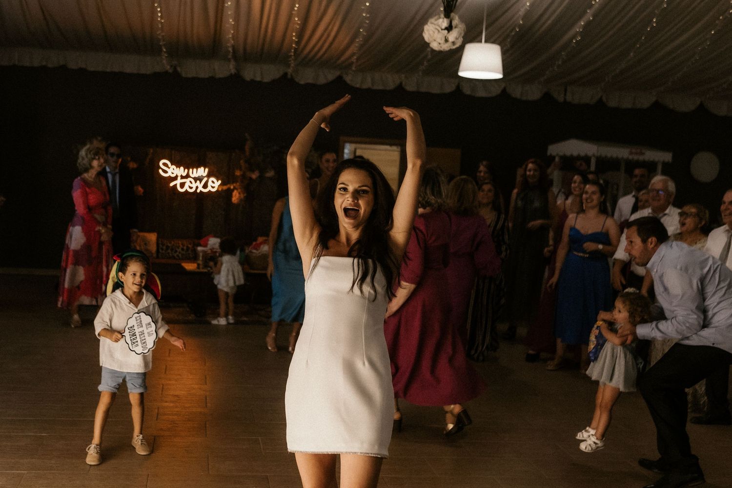 Wedding guests dancing and celebrating on a dimly lit dance floor at an indoor reception venue.