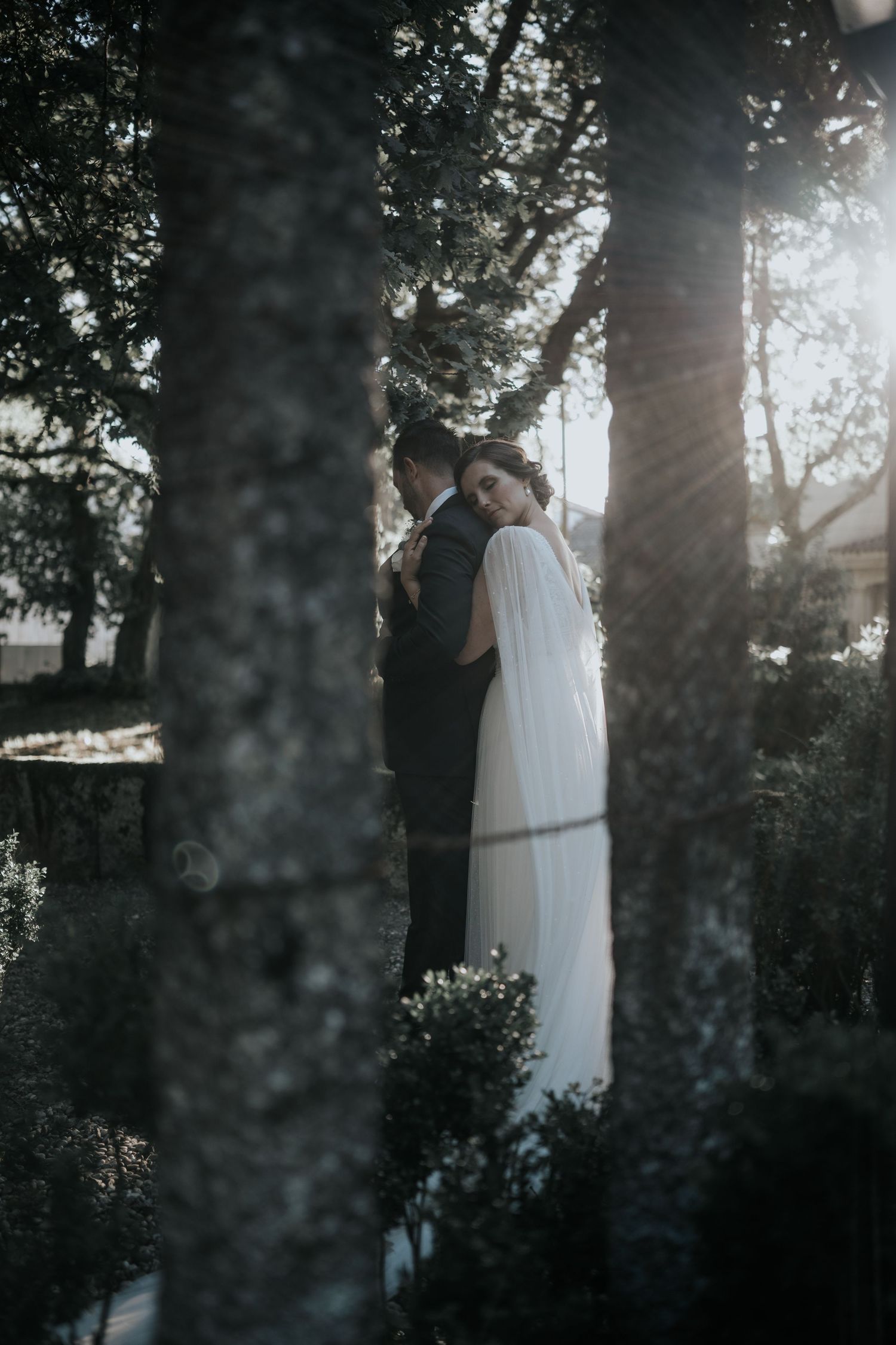 An artistic wedding photo through trees showing a couple in white dress and dark suit in dreamy lighting.