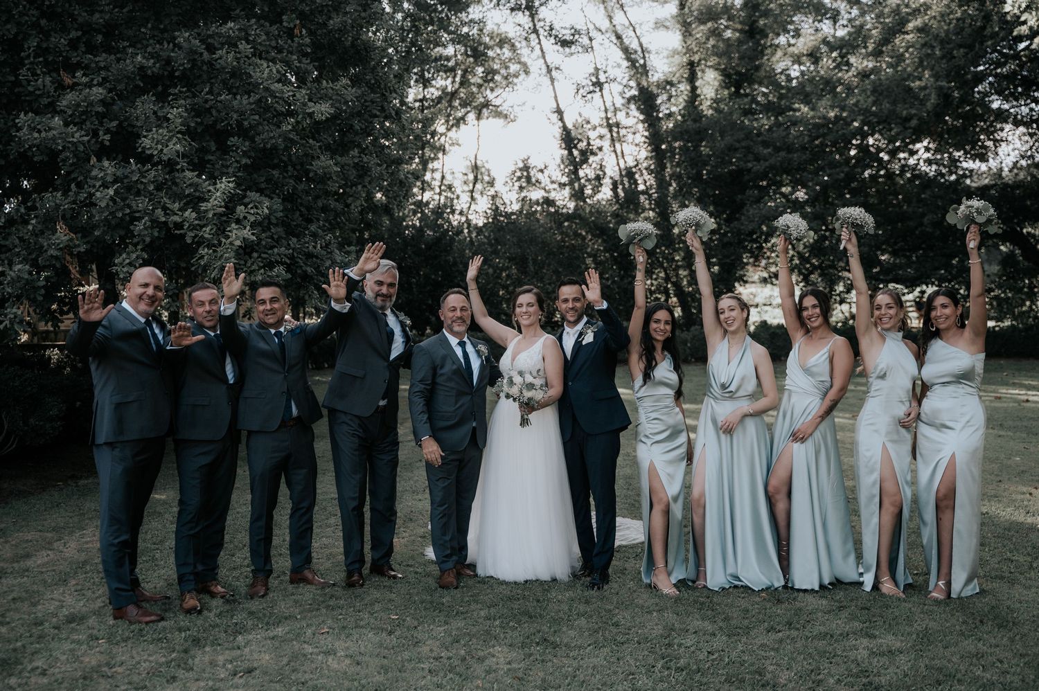 Wedding party poses together outdoors with bridesmaids in light dresses and groomsmen in dark suits.