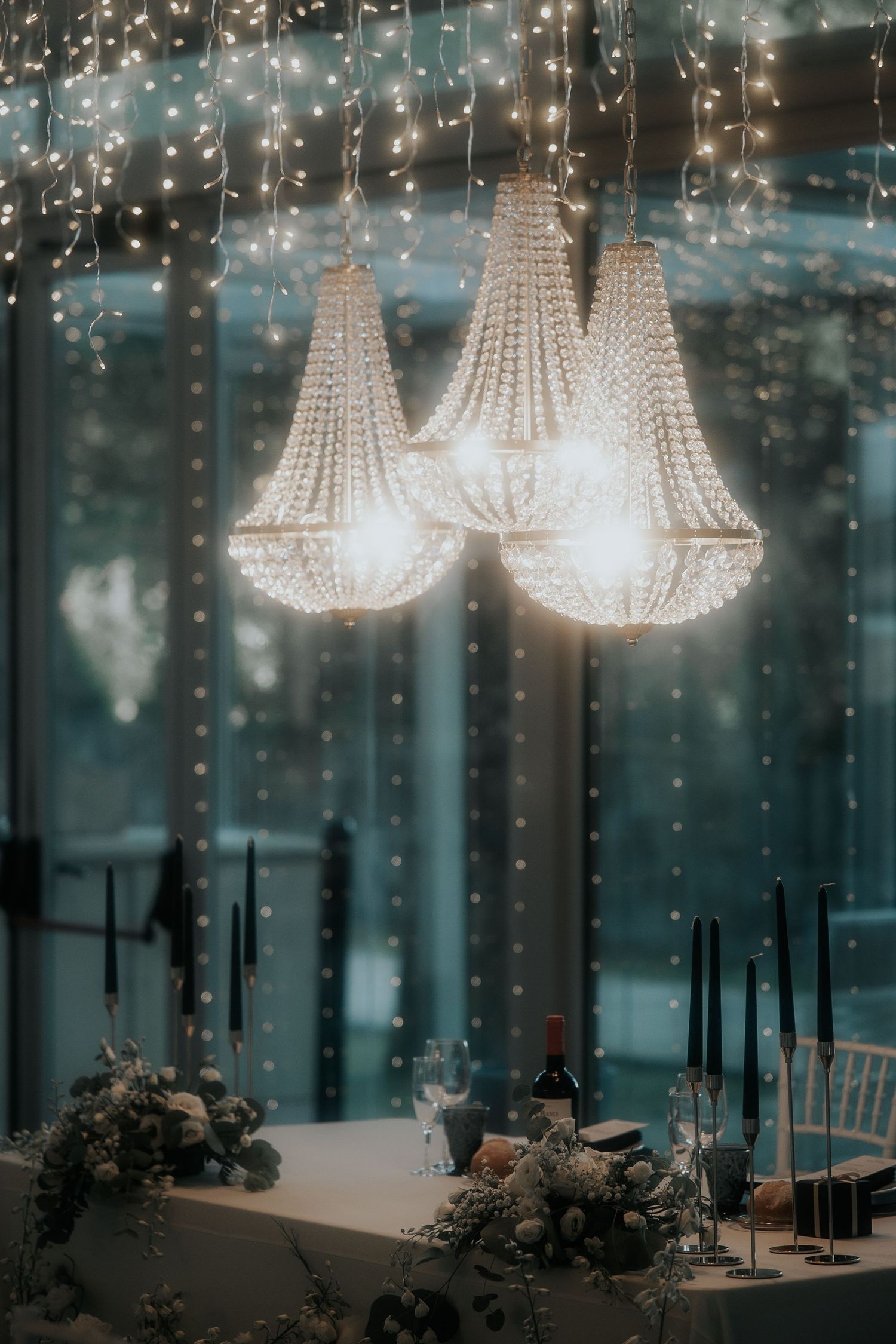 Crystal chandeliers and string lights create an elegant ambiance in a dimly lit reception venue with glass walls.