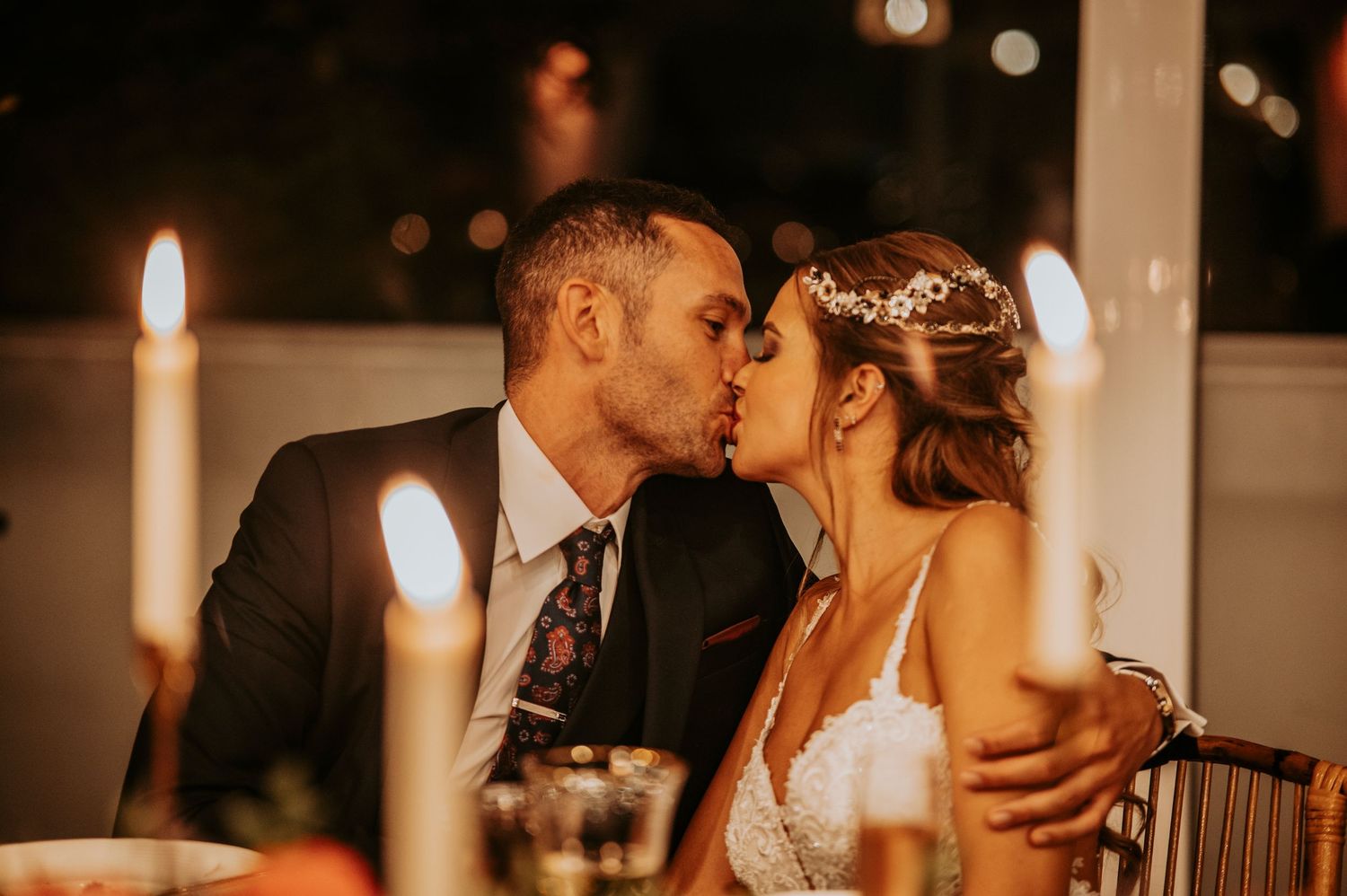 A romantic wedding couple shares an intimate kiss at a candlelit reception dinner table.